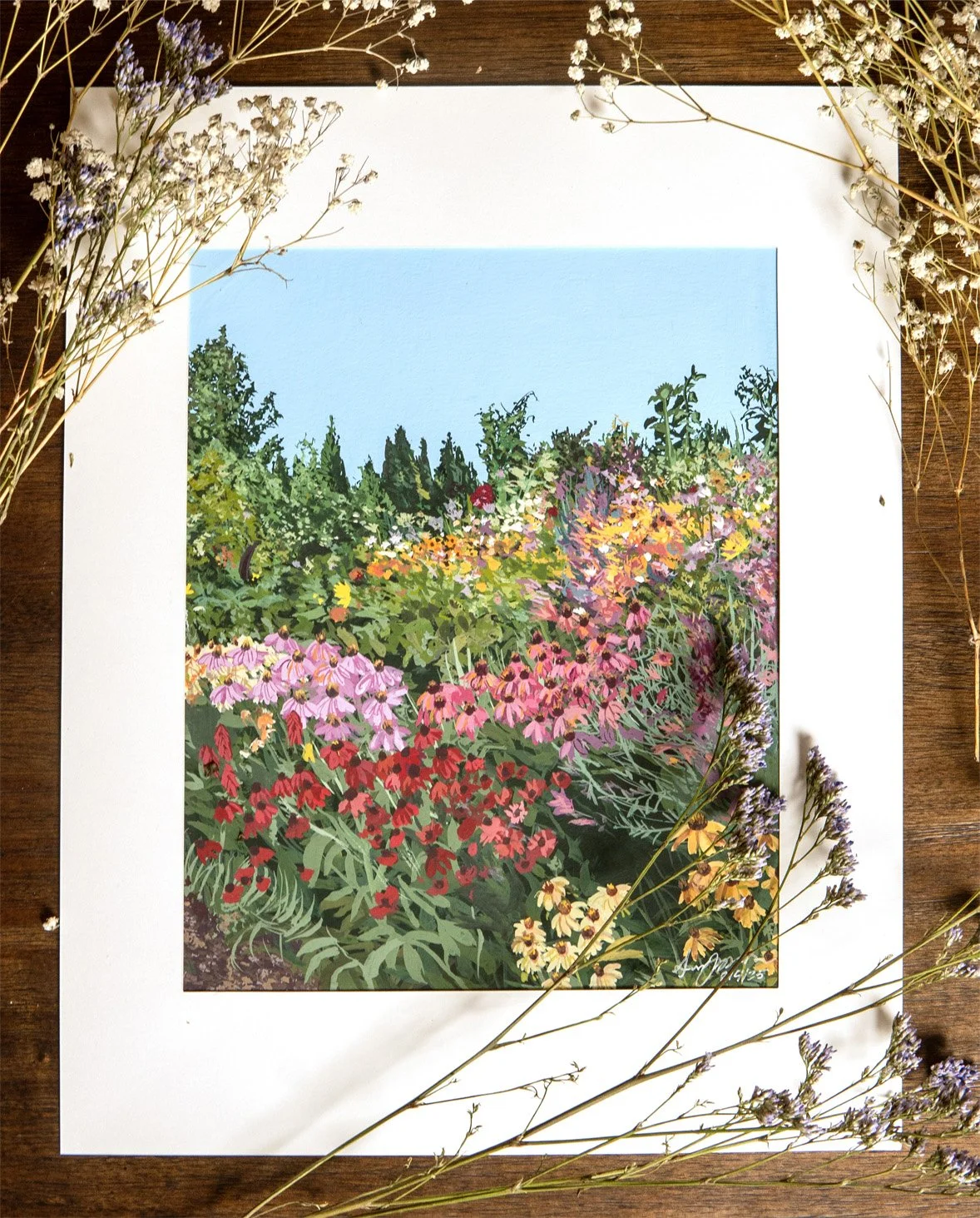 A painting of a colorful garden with pink, red, yellow, and purple flowers under a bright blue sky, surrounded by sprigs of dried flowers on a wooden surface.