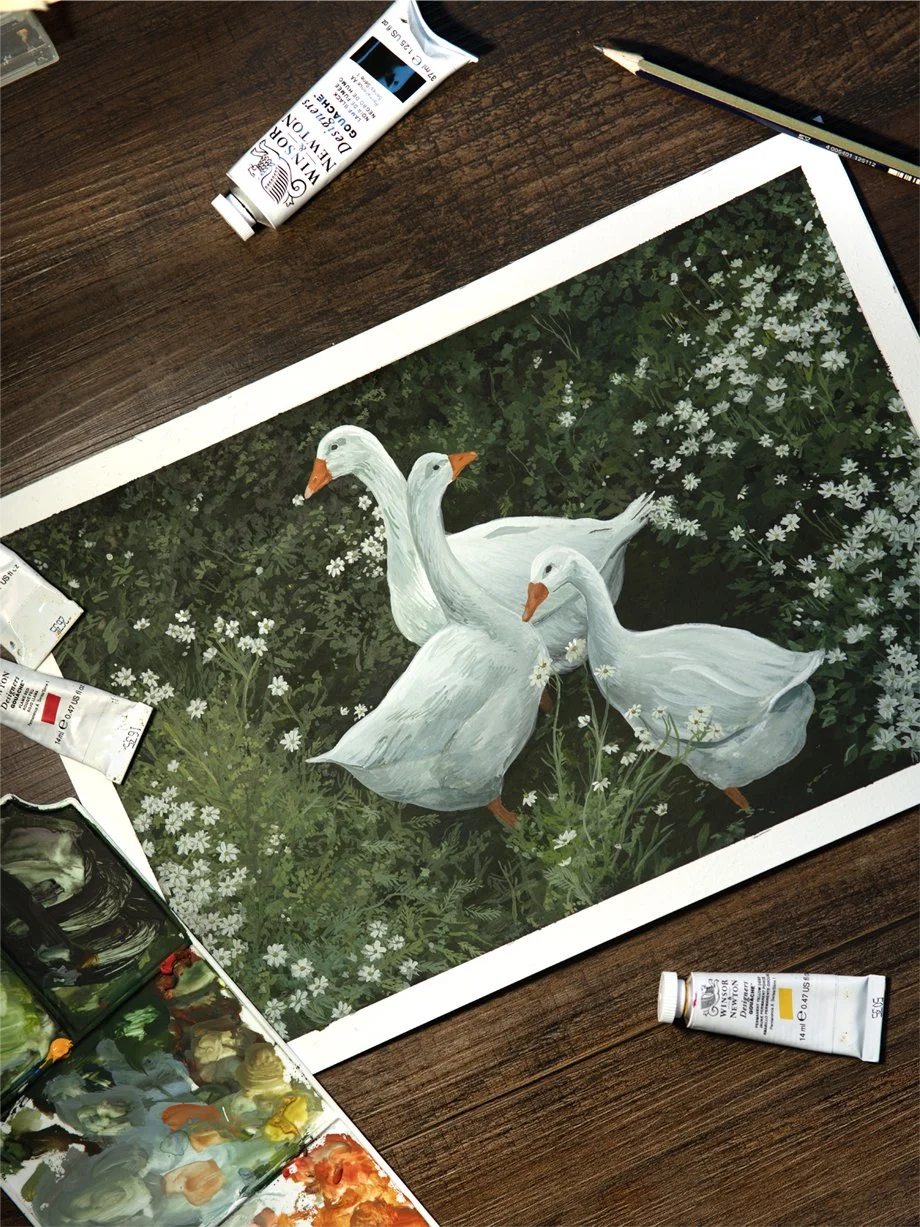 Painting of three white geese in a green field with flowers, on a wooden table with tubes of paint, a palette, a pencil, and paper.