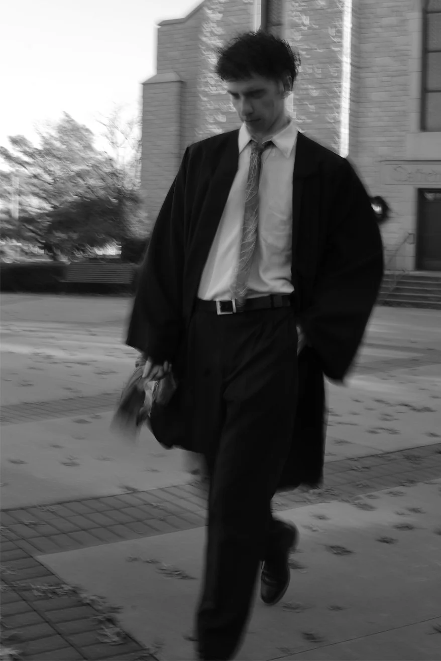 A young man in formal attire, including a suit, tie, and dress shoes, is walking outdoors near a building with stairs. The photo is in black and white.
