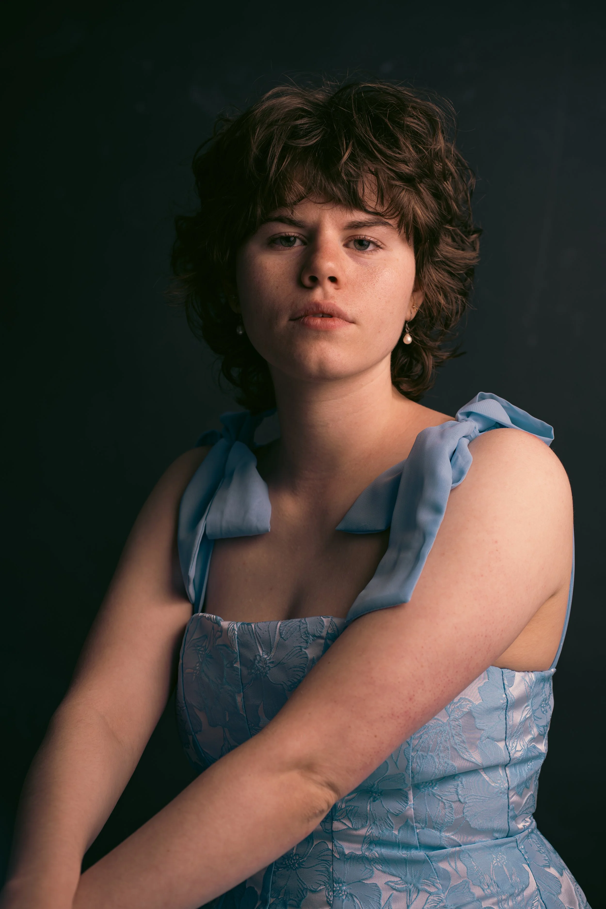 Portrait of a young woman with short, curly brown hair wearing a light blue dress with bows on the shoulders and pearl earrings against a dark background.