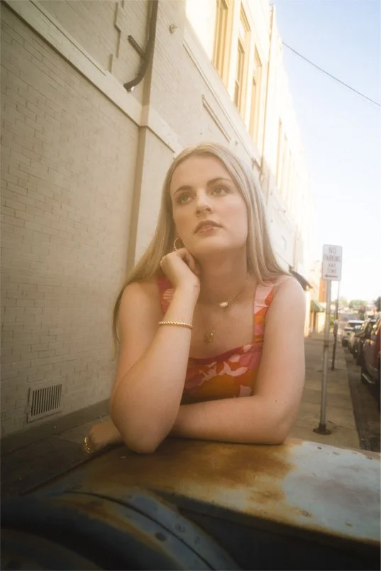 A young woman with blonde hair sitting at an outdoor cafe table, leaning on her hand, looking thoughtful, with a beige building and parked cars in the background.