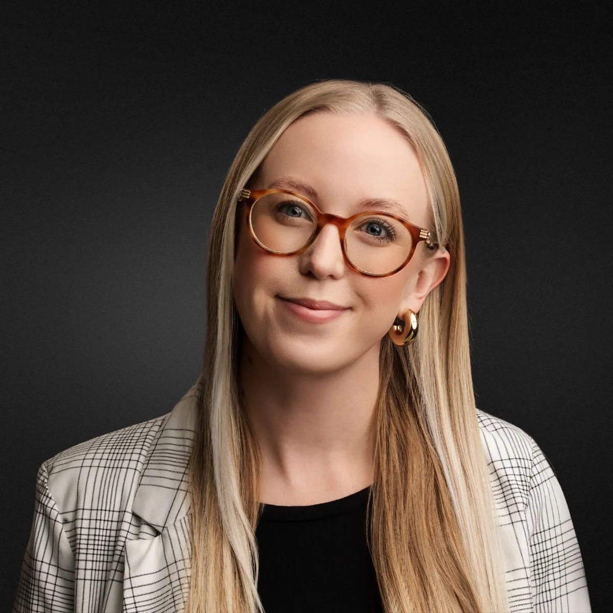 Portrait of a young woman with blonde hair, wearing large tortoise shell glasses, gold hoop earrings, a black top, and a checkered blazer, against a dark background.