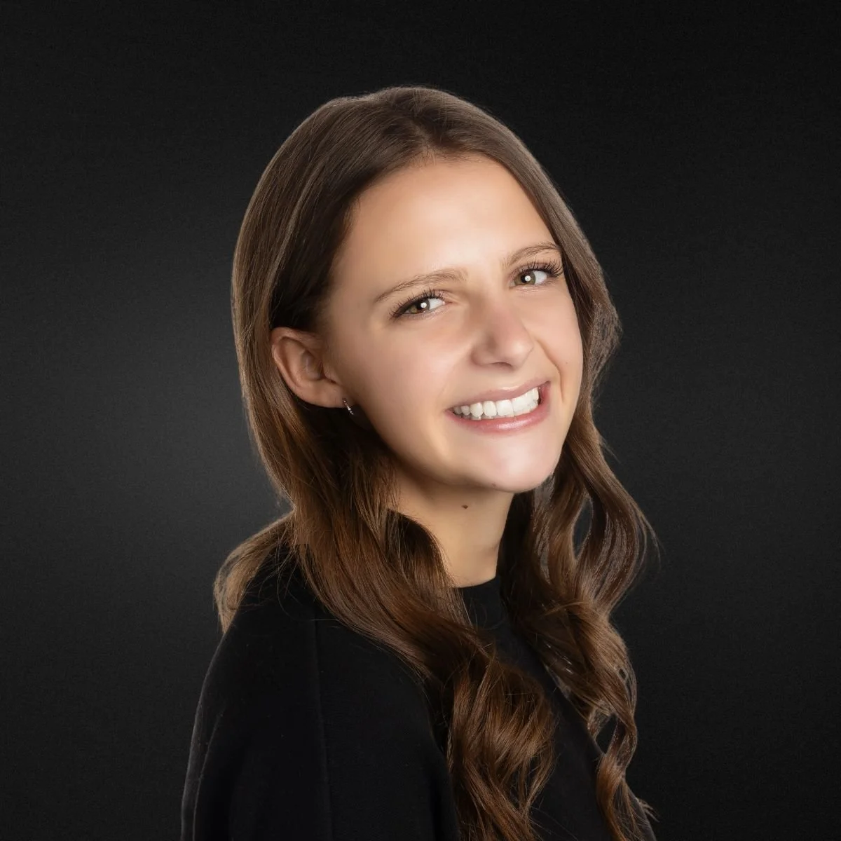 Portrait of a young woman with long brown hair, smiling, against a dark background.