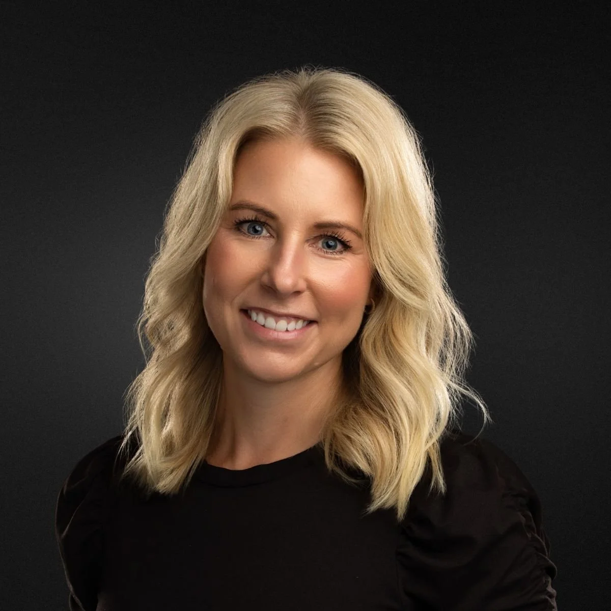 Portrait of a blonde woman with wavy hair, smiling, wearing a black top, against a dark background.