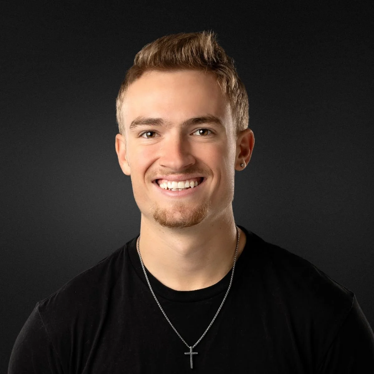Portrait of a young man smiling, wearing a black shirt, a cross necklace, and an earring, against a dark background.