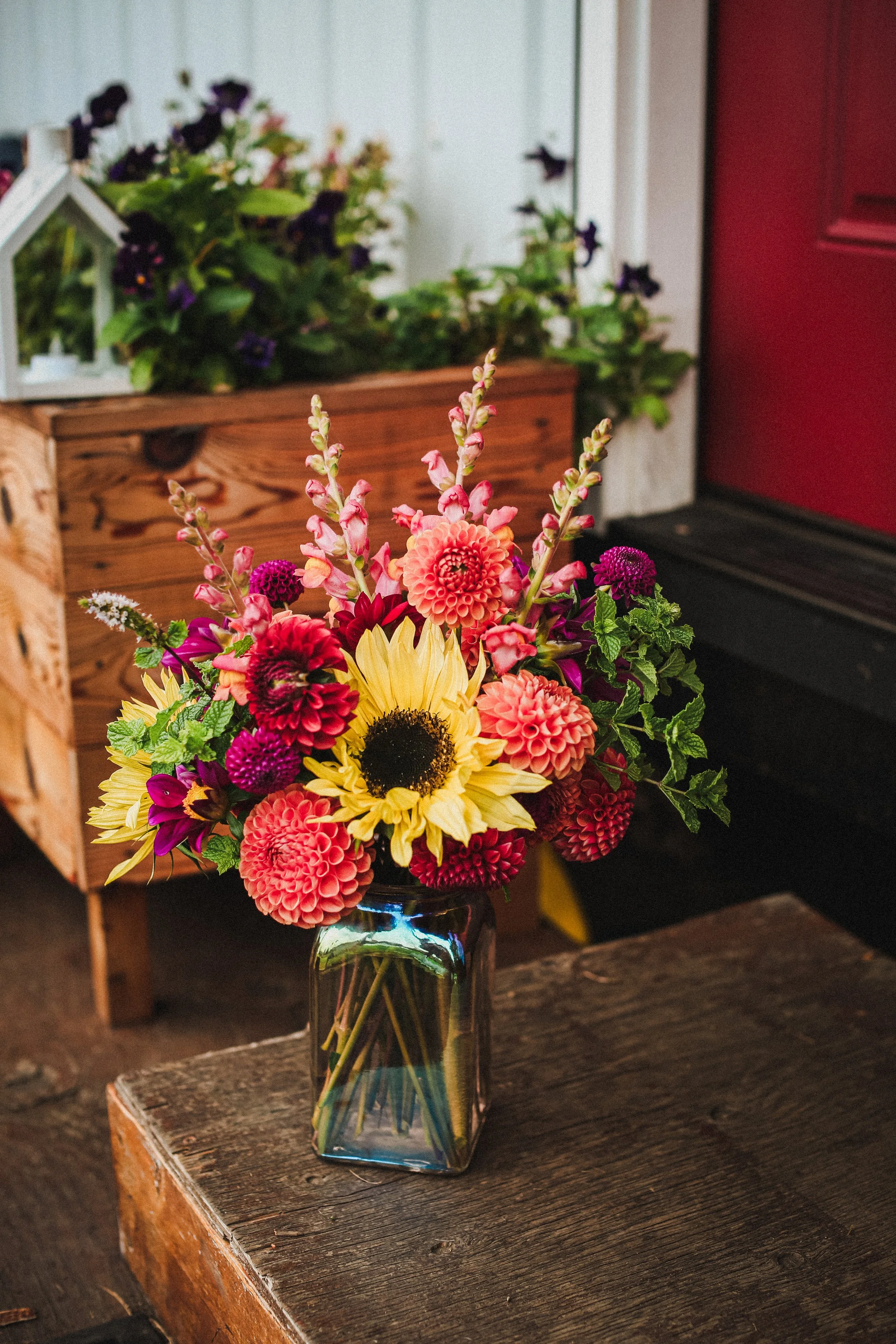 A colorful bouquet of flowers including sunflowers, dahlias, and snapdragons in a glass vase on a wooden table near an entryway.
