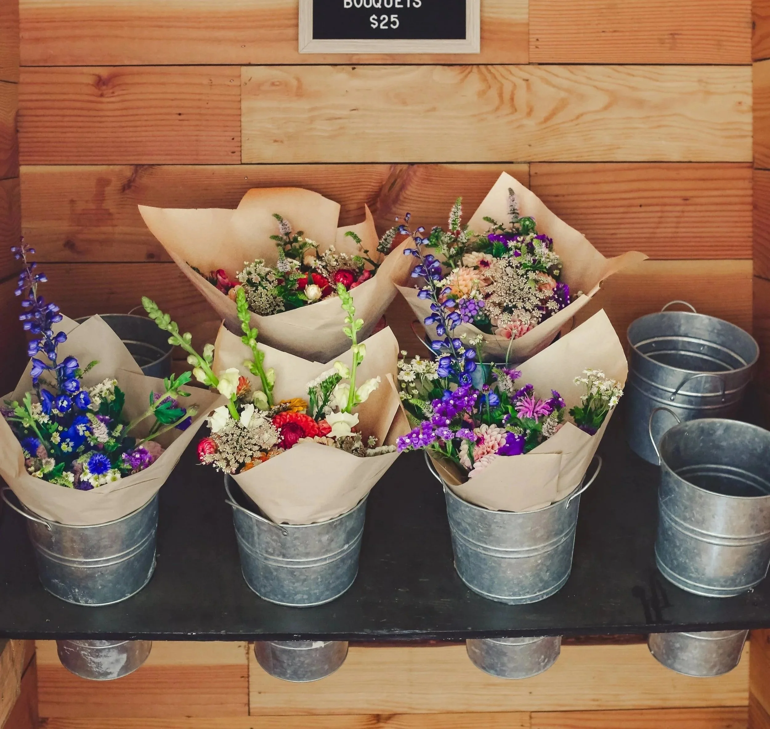 Multiple bouquets of mixed flowers wrapped in paper inside metal buckets on a black surface, with a wooden wall in the background and a sign reading 'Bouquets".
