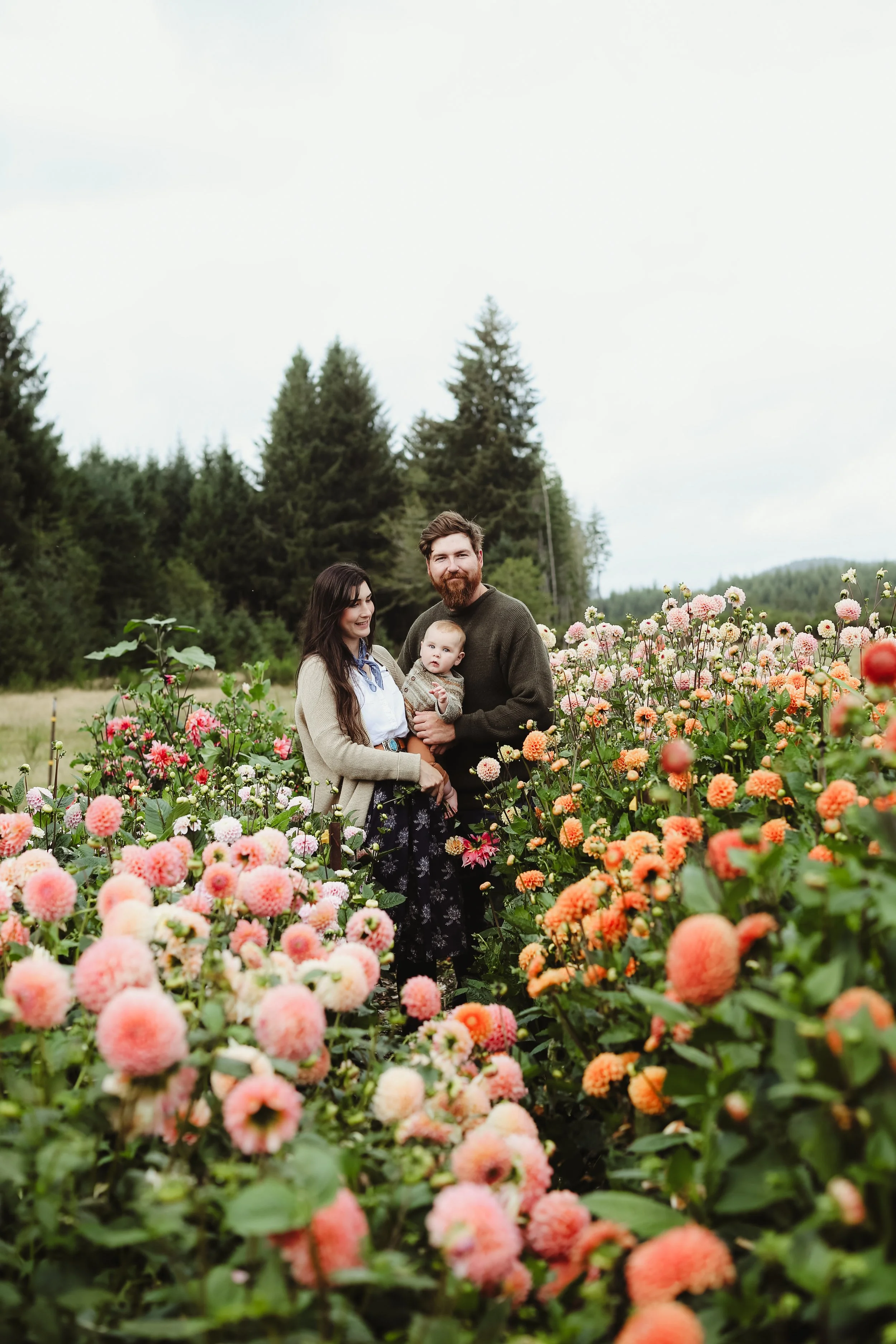 A family of three standing in a colorful flower garden with pink, orange, and white blooms, surrounded by green trees and forest in the background.