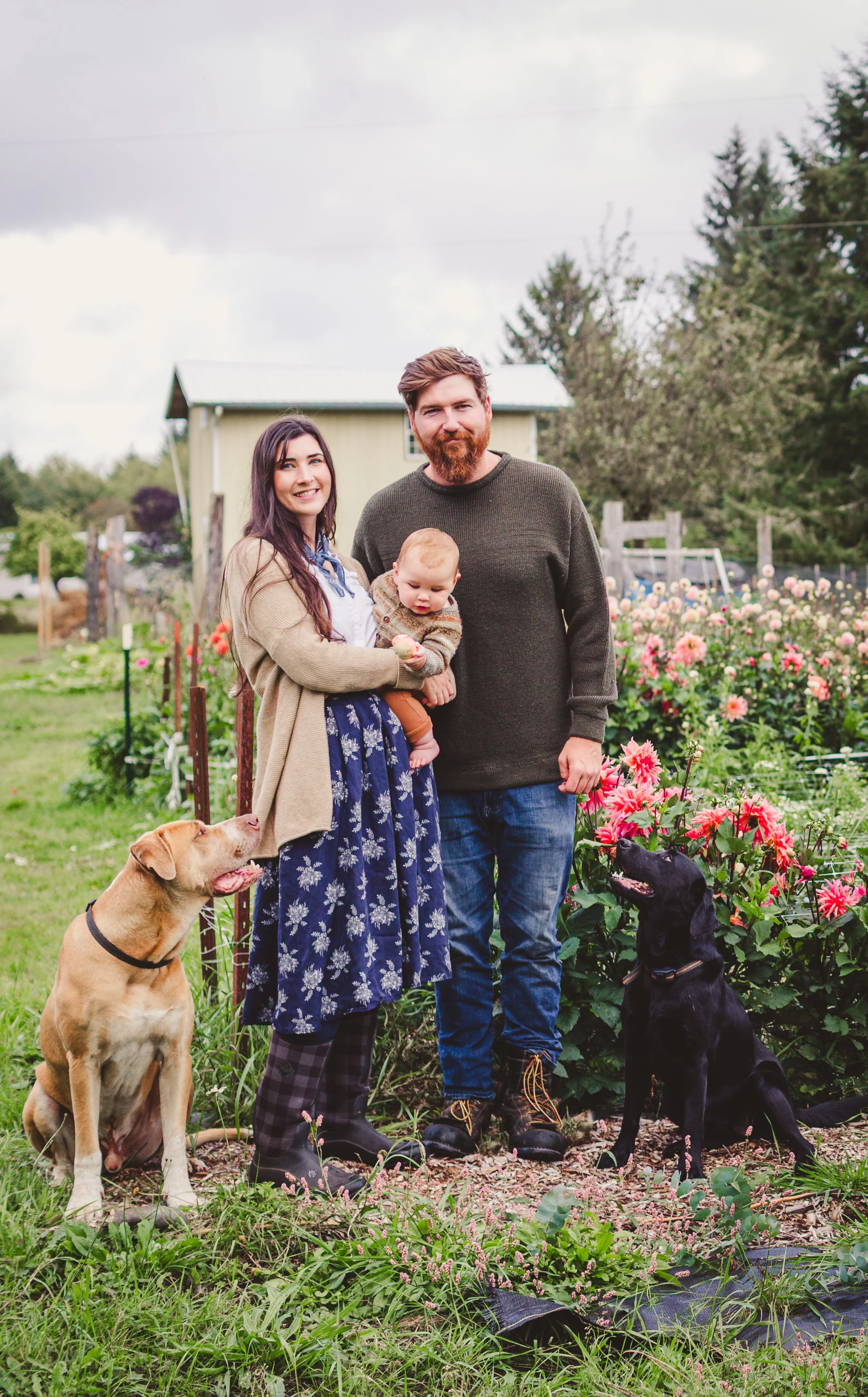 A family of three with two dogs standing outdoors in a garden filled with blooming flowers and greenery, with a small shed and trees in the background.