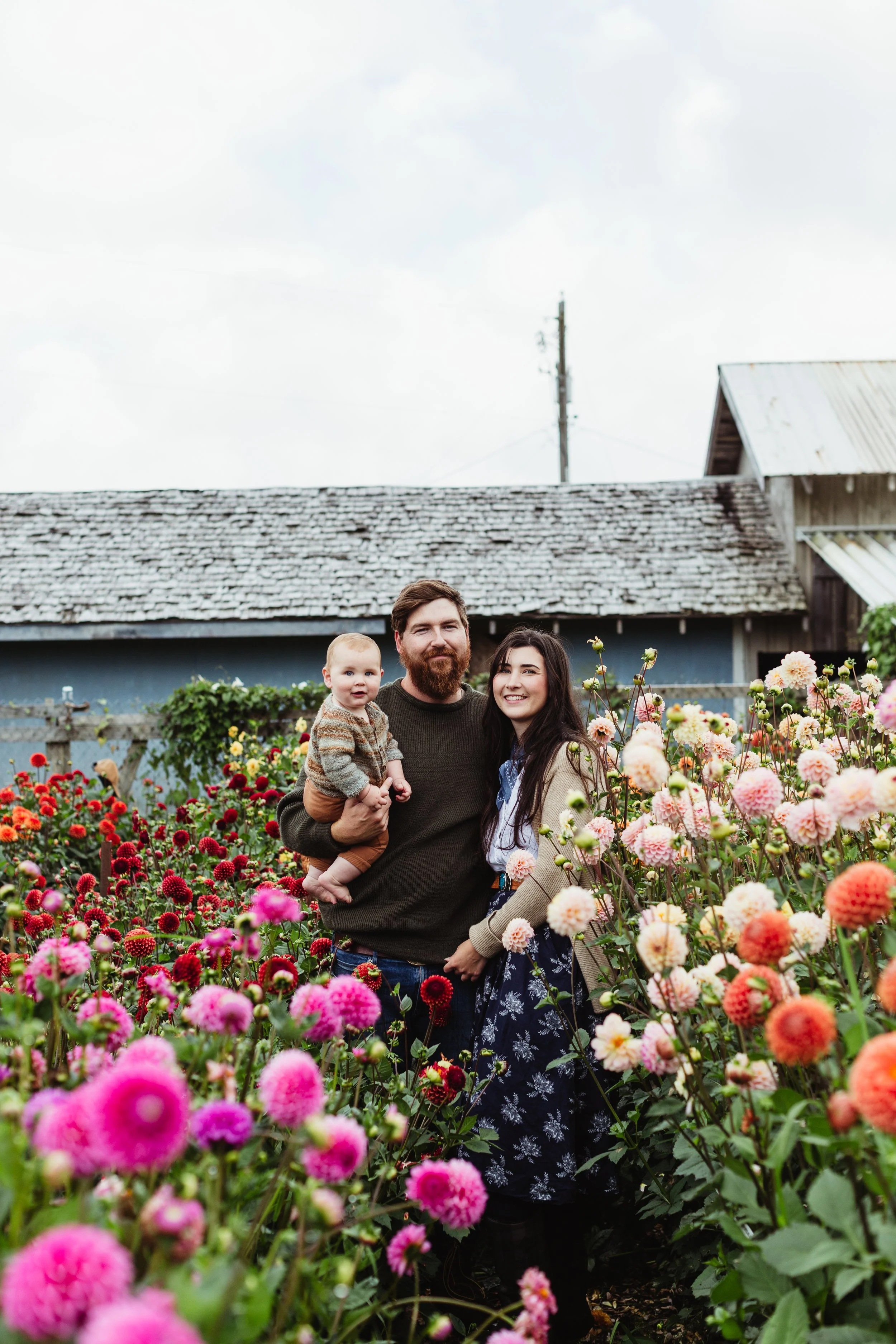 A family of three standing in a colorful flower garden with a barn and an overcast sky in the background.