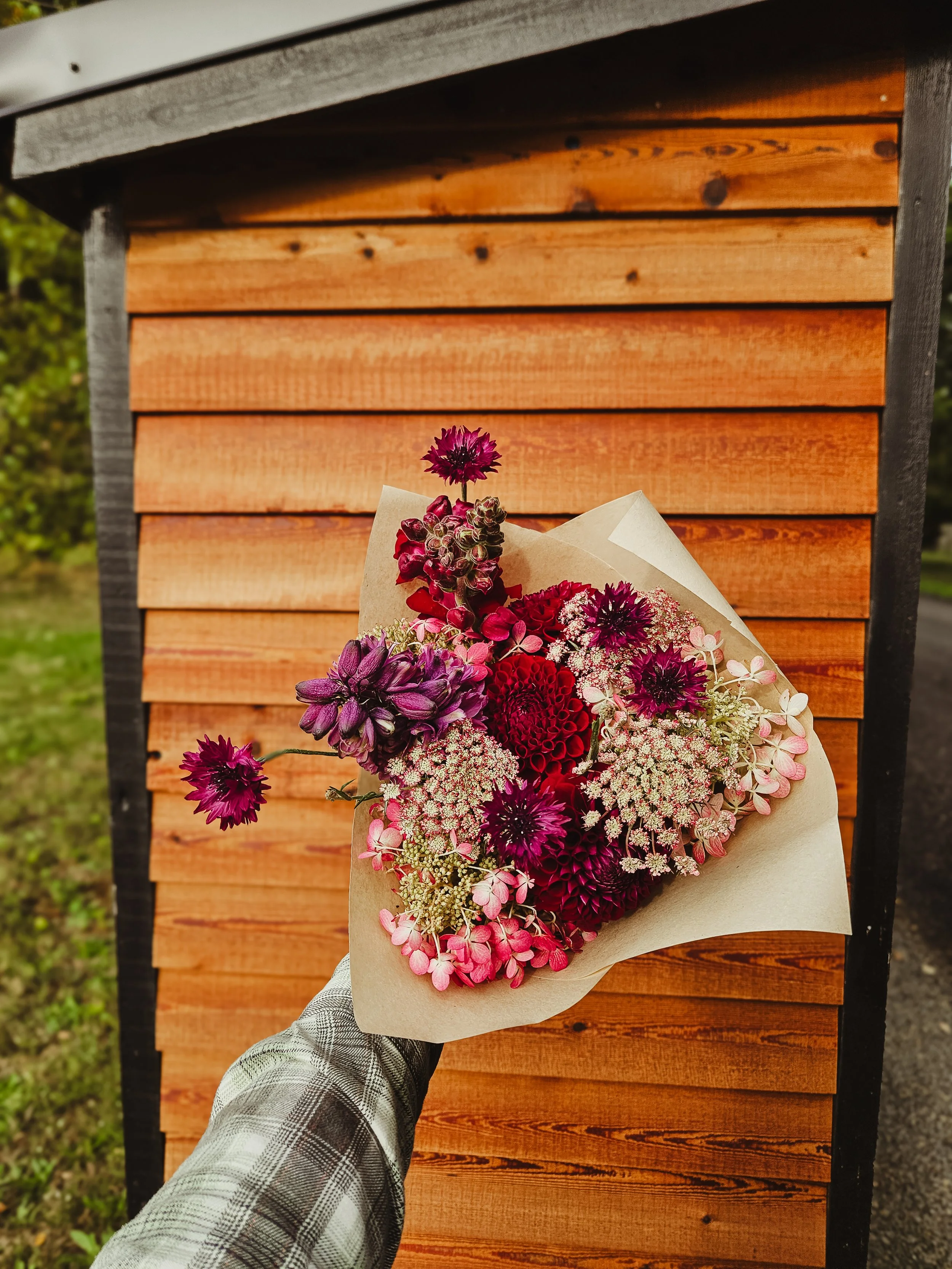 A person's hand holding a bouquet of pink, purple, and white flowers in front of a wooden structure.
