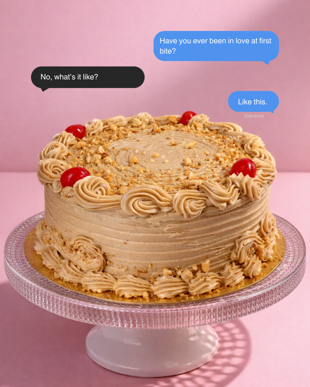 A layered cake with beige frosting, red cherries, and chopped nuts on top, displayed on a glass cake stand.