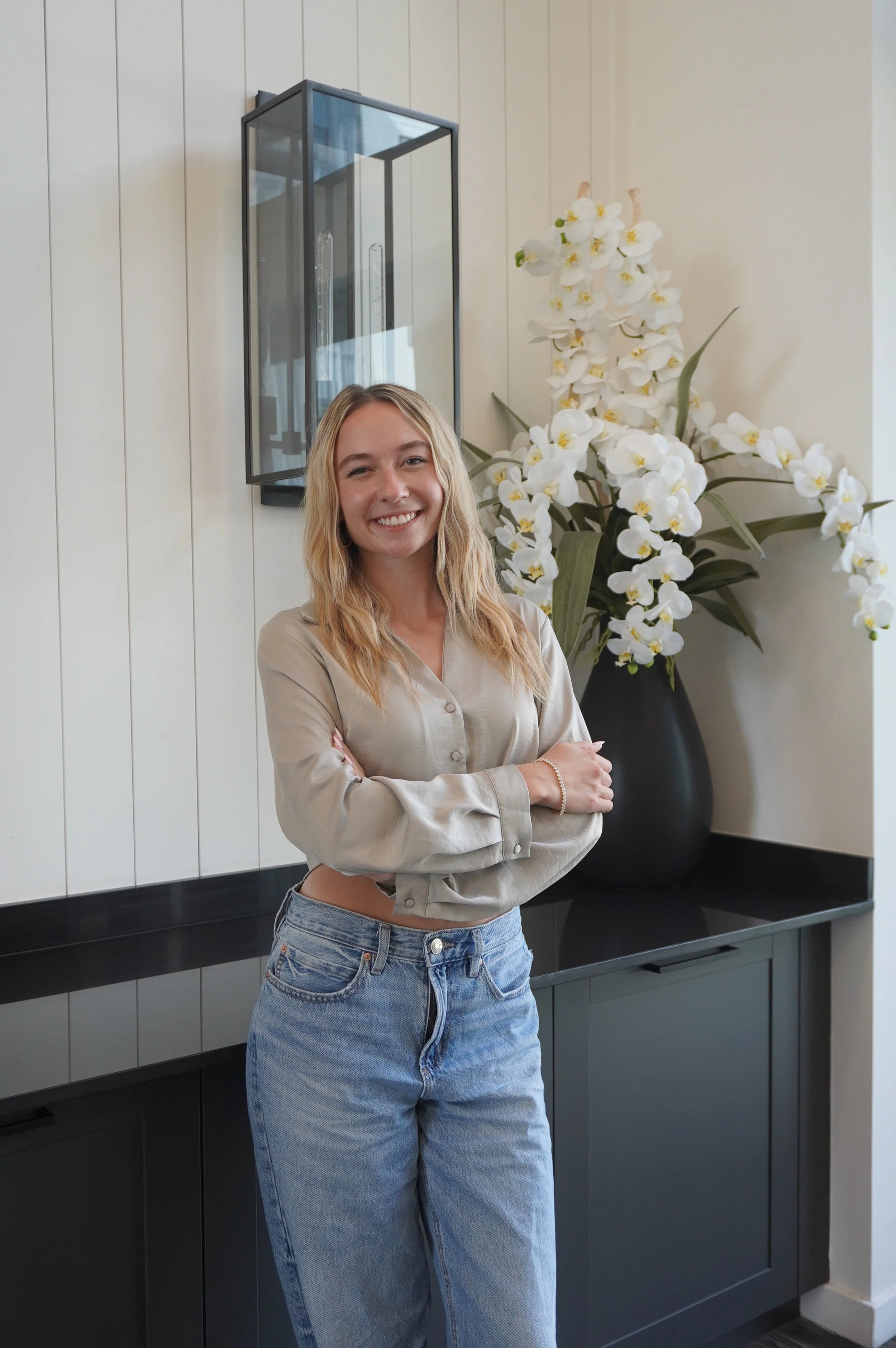 A smiling young woman with blonde hair standing indoors next to a large black vase filled with white orchids, against a cream-colored wall.