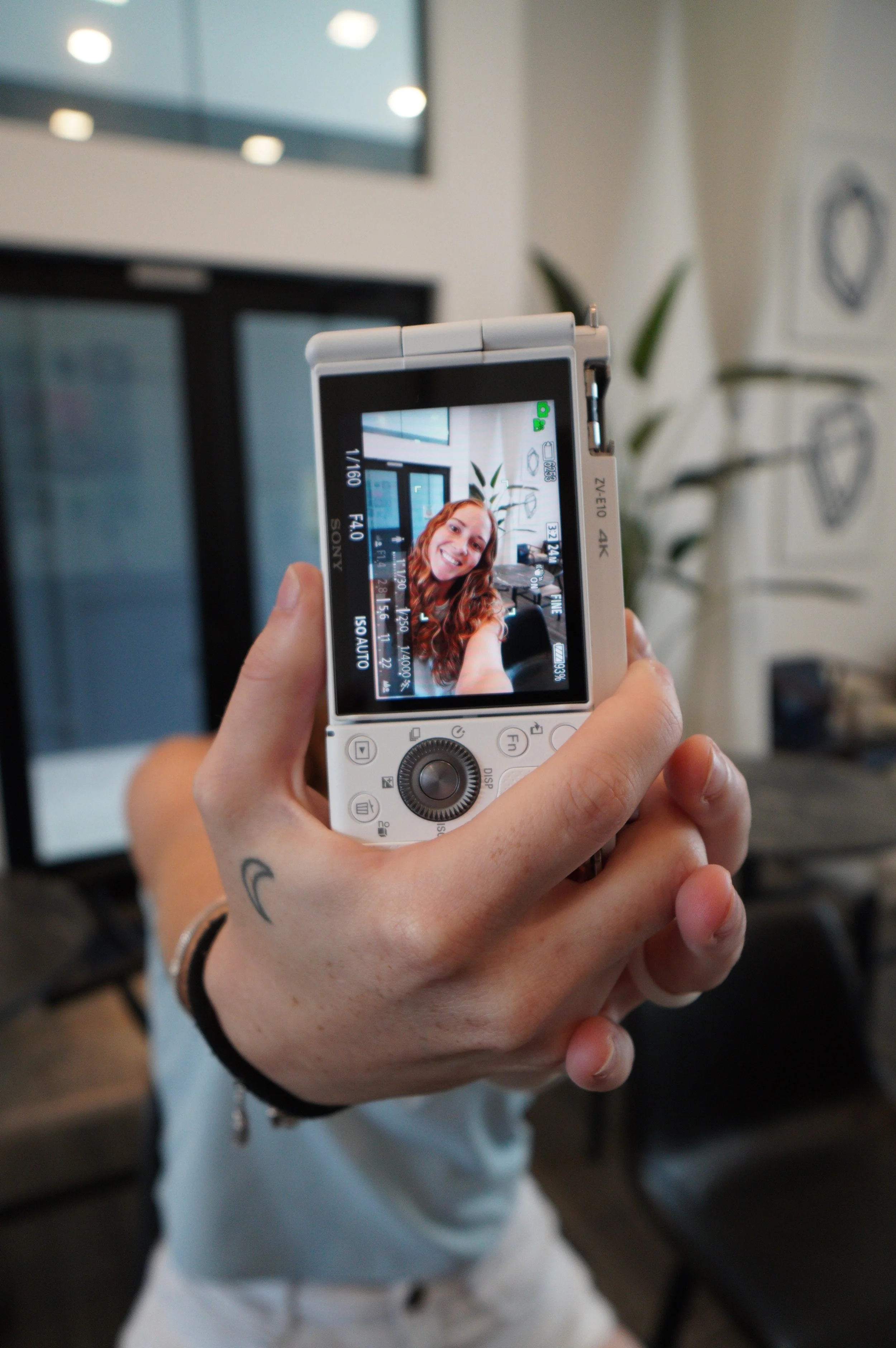 Person taking a selfie using a compact digital camera in a modern indoor setting.