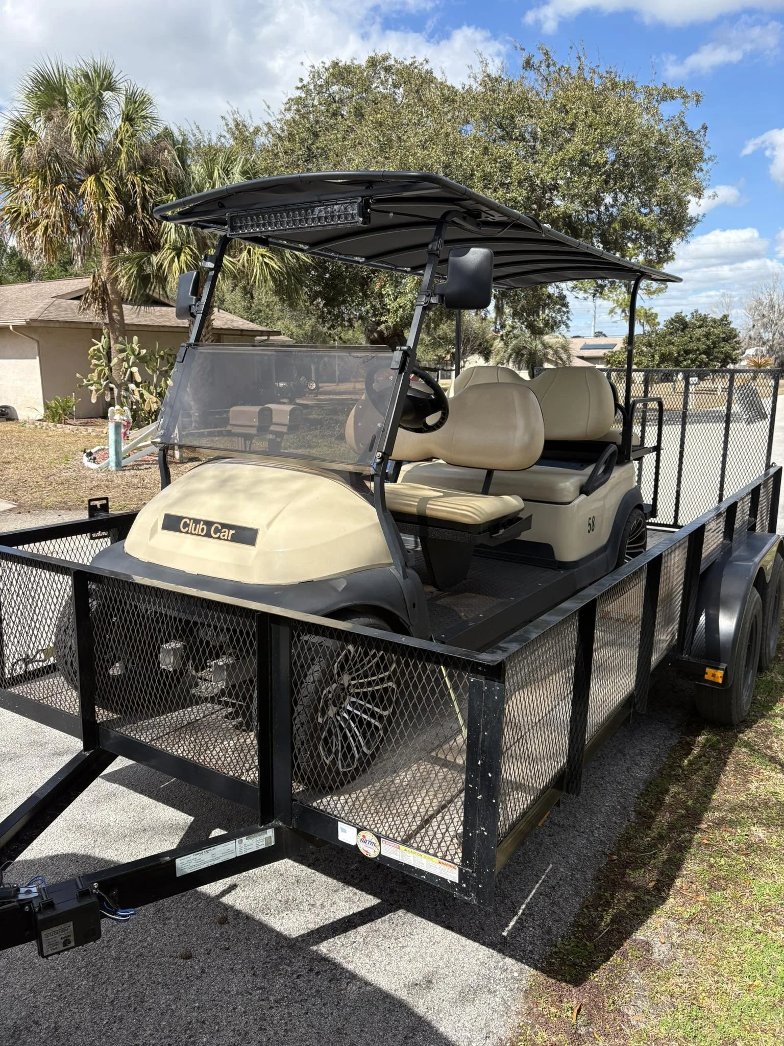 A beige golf cart with a black roof parked on a trailer, with a front windshield and beige seats, surrounded by a fenced yard with trees and a house in the background.