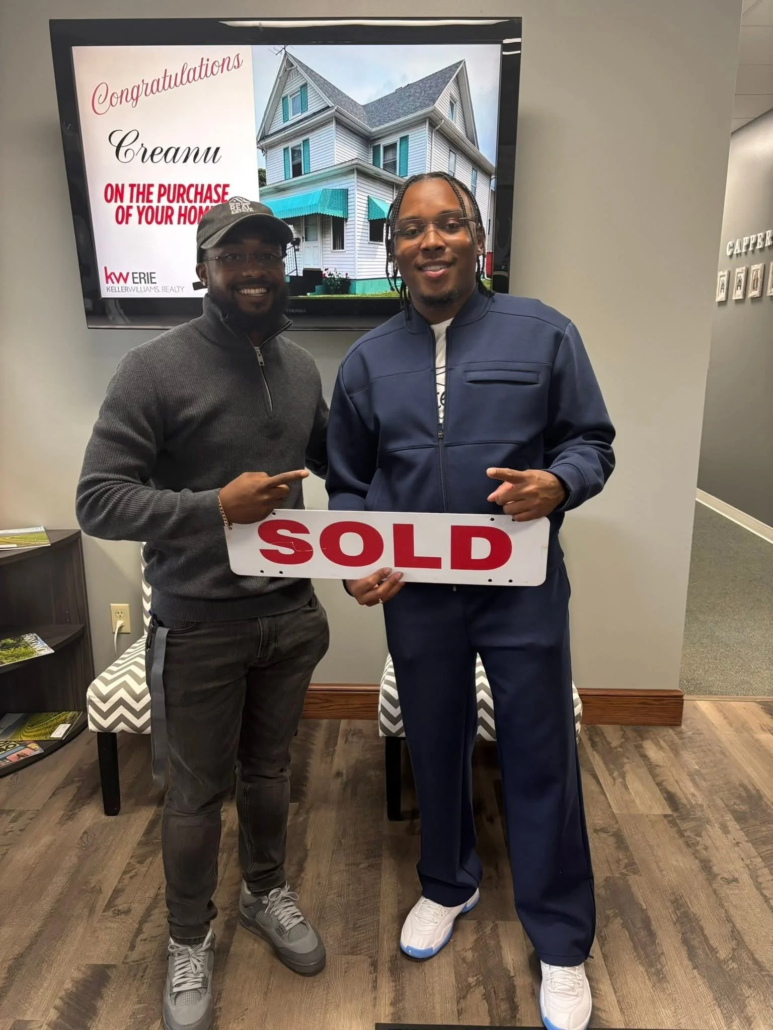 Two men smiling and holding a 'SOLD' sign, standing in front of a screen that reads, 'Congratulations!'