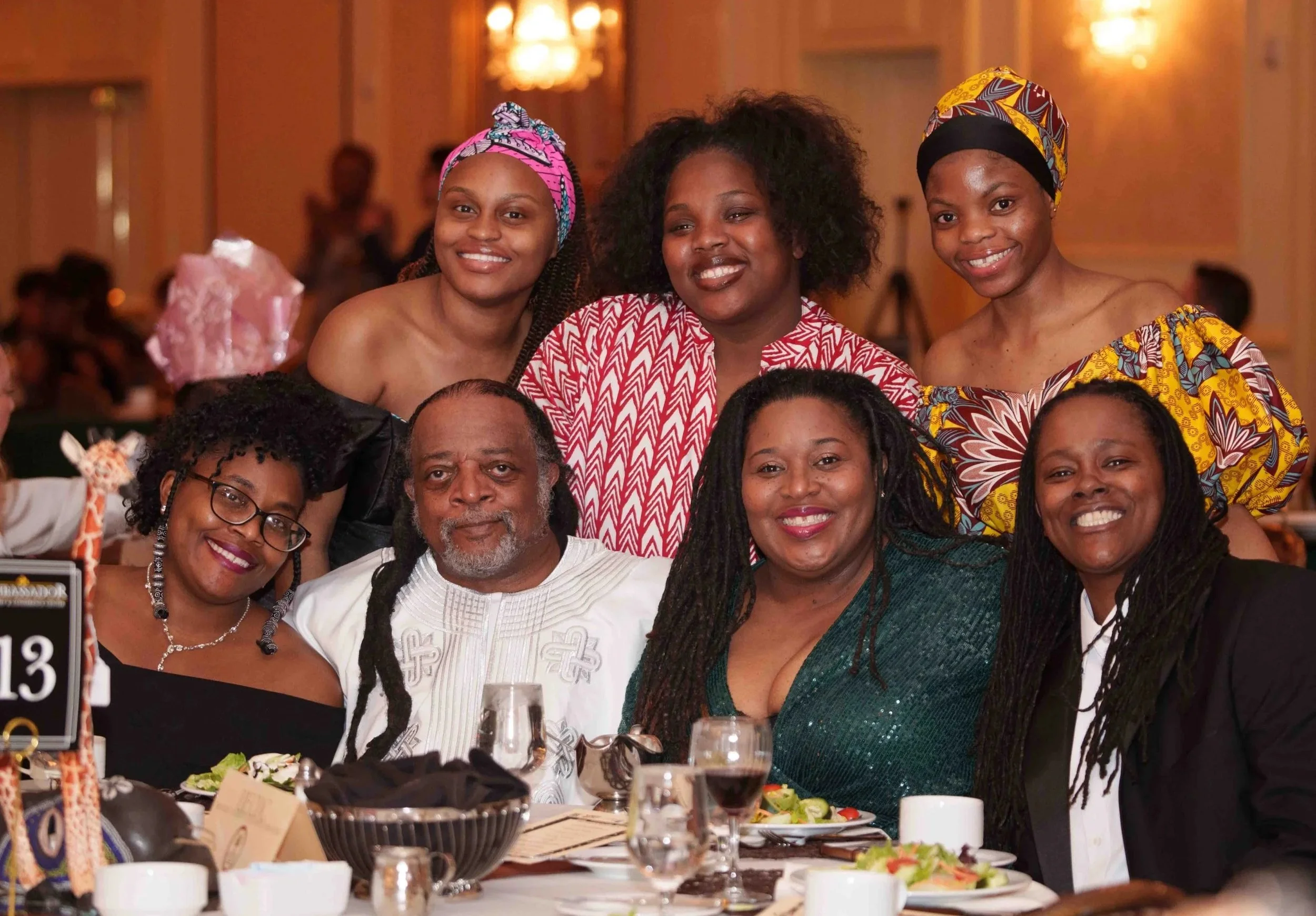 Group of people dressed in formal wear and authentic African apparel, smiling while sitting around a table.