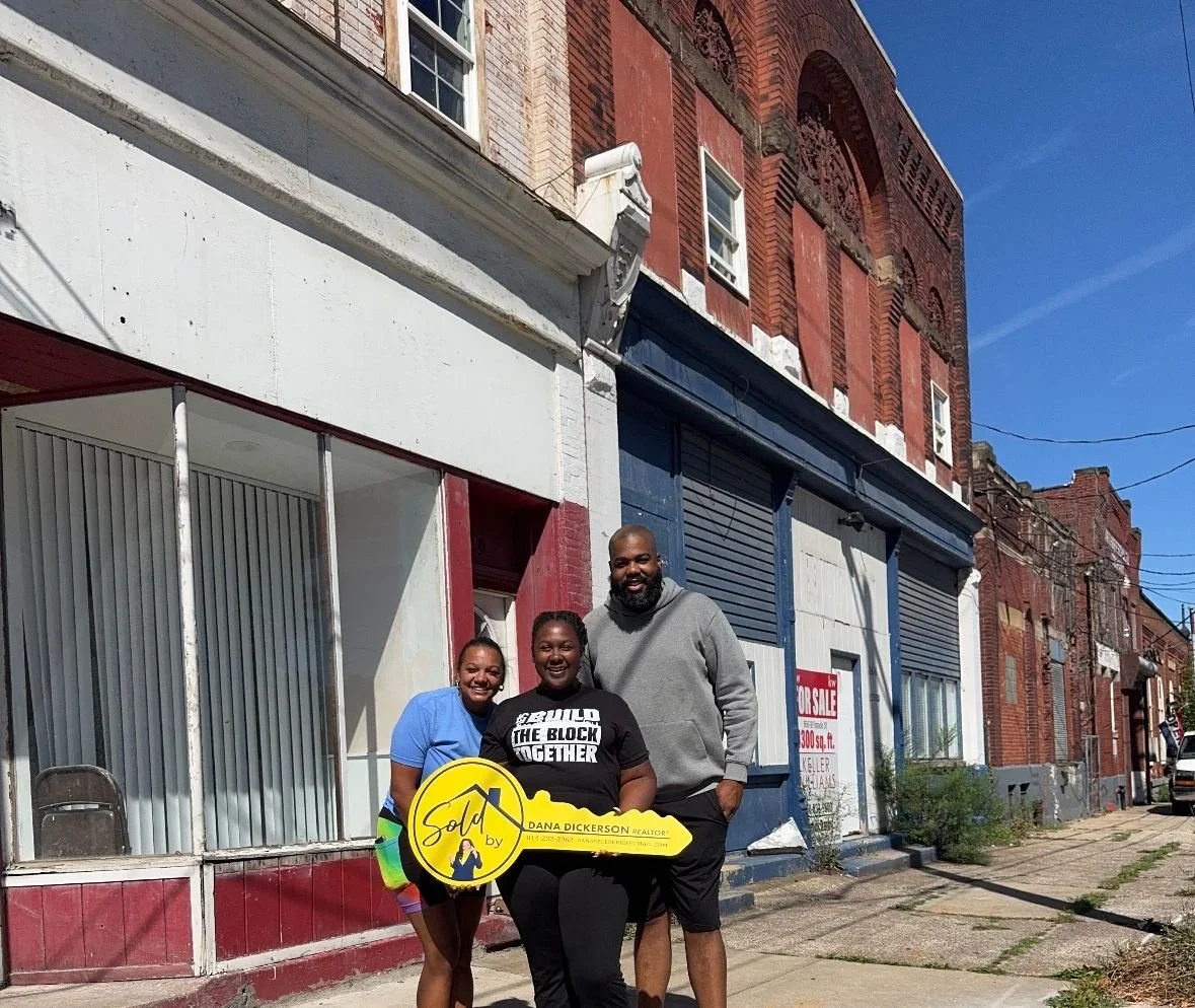 Three people smiling, standing outside two old buildings, holding a large key that says, 'SOLD.'