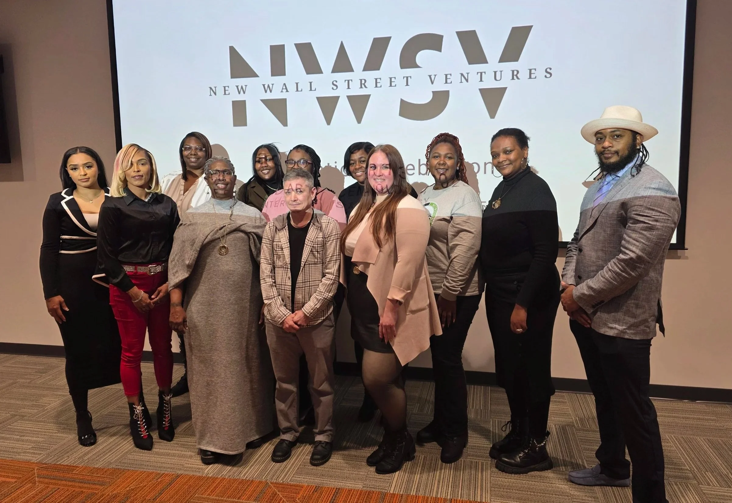 Group of people of various races, smiling, standing in front of a large projector screen with 'New Wall Street Ventures' on it.