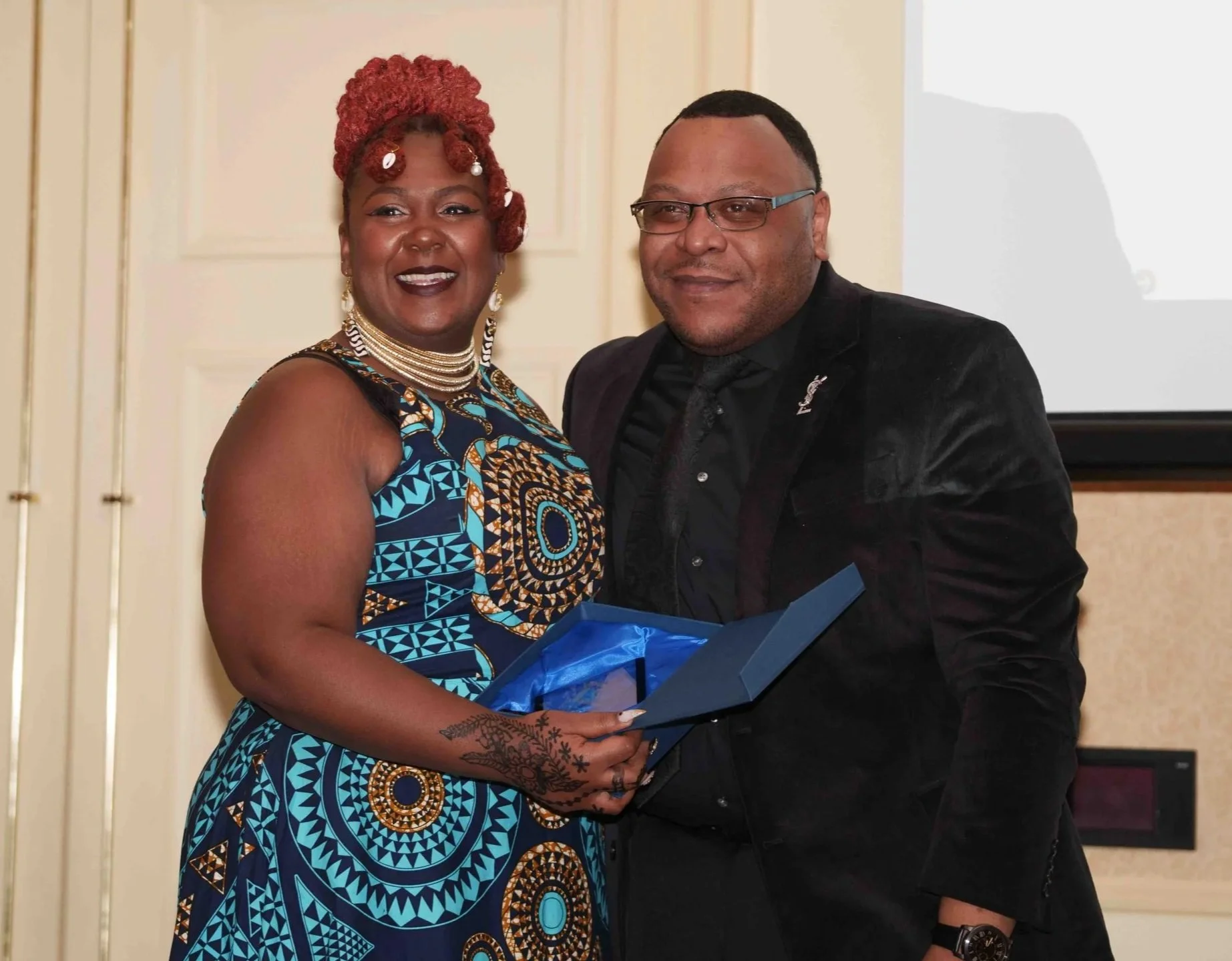 A man and woman smiling, while the woman hands a glass award to the man.