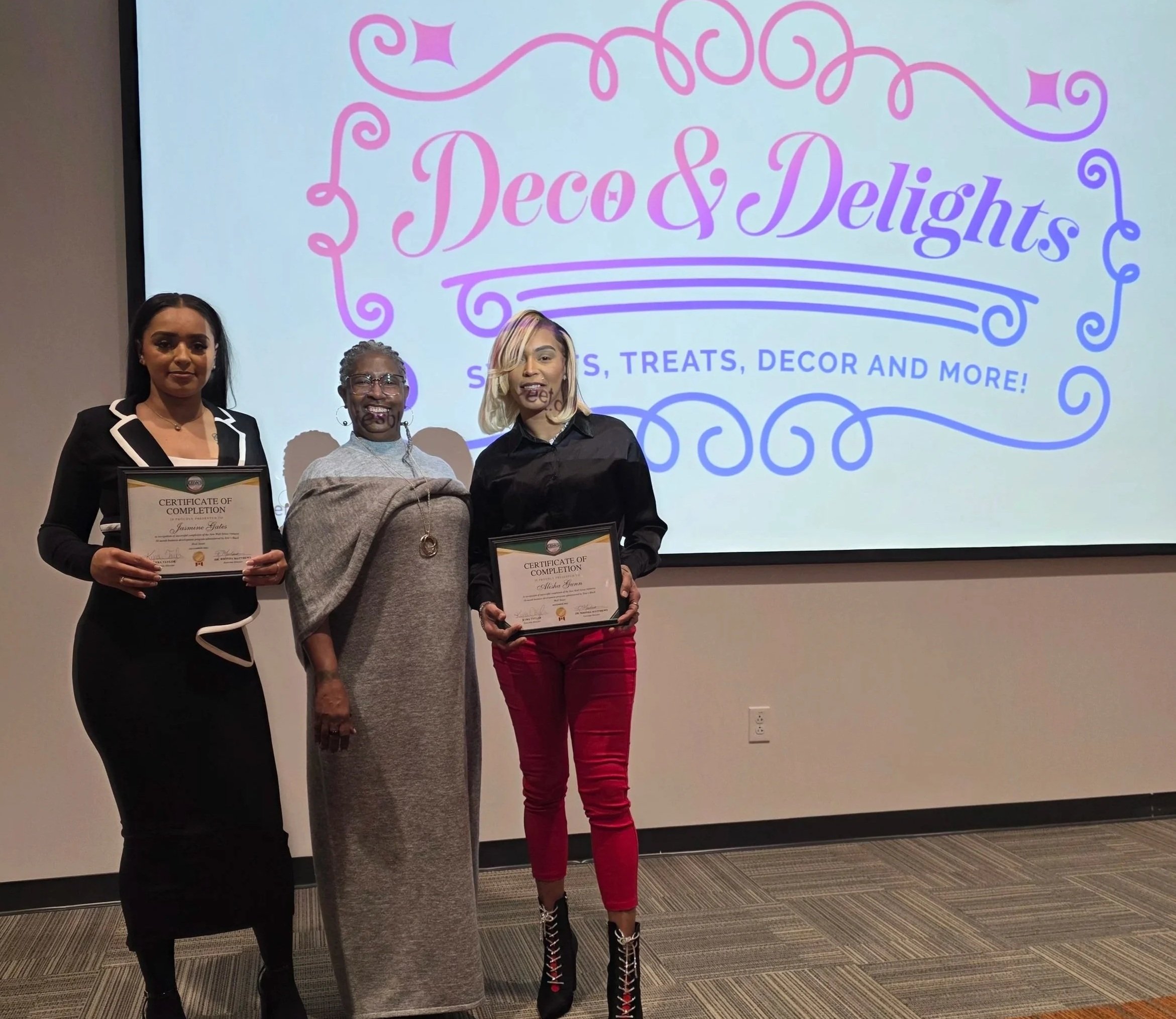 Three women smiling, two holding certificates. Infront of a large screen that says, 'Deco & Delights.'