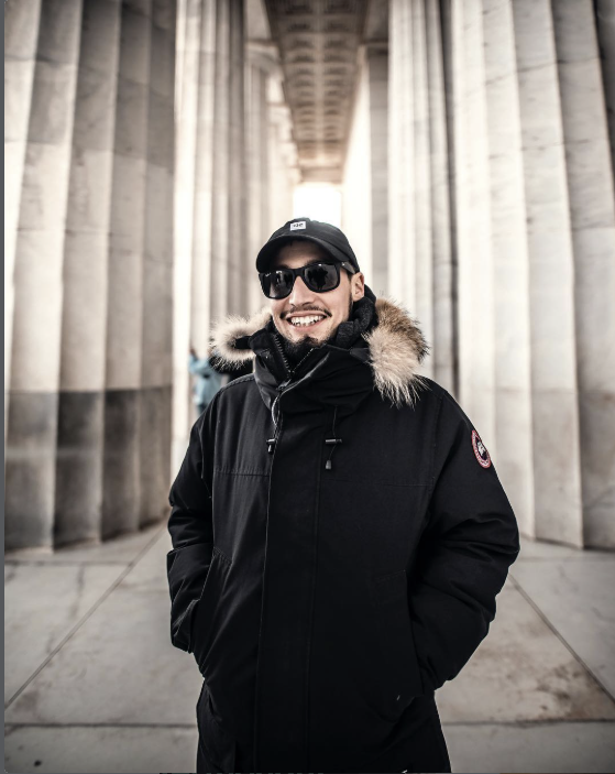A smiling man wearing sunglasses, a black cap, and a black winter coat with a fur-lined hood, standing between large stone columns at a historic site.
