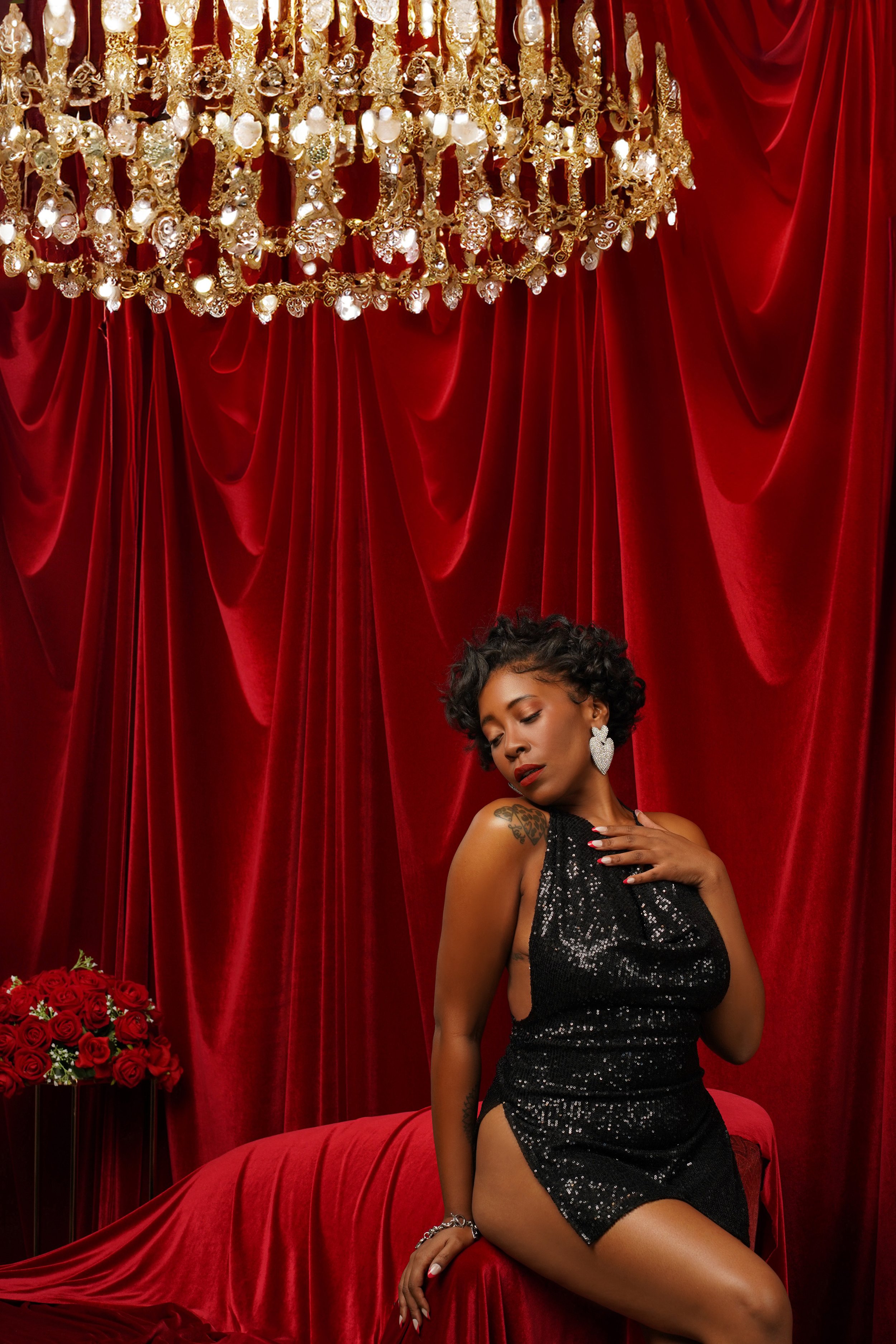 black woman slightly sitting on the edge of the couch clutching her chest slightly. red couch with red velvet curtains. gold chandelier hanging. Her eyes are closed with beautiful black curly hair wearing heart diamond earrings from fashion nova.