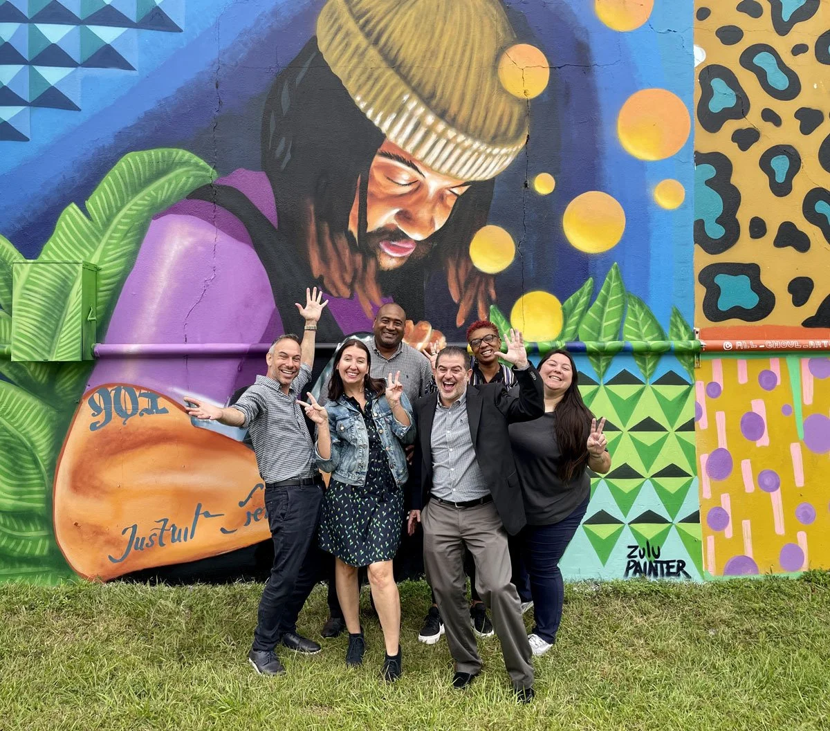 Group of six smiling people, standing in front of a colorful mural of a woman with dreadlocks and a beanie, surrounded by vibrant geometric and animal-print patterns