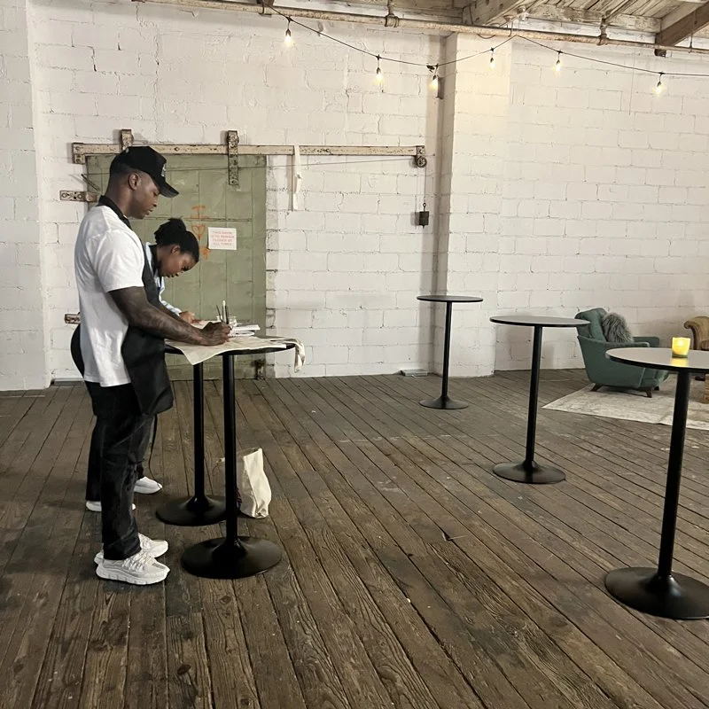 Two people standing at a high table in a minimalistic, industrial-style room with wooden floors and a white brick wall, where a green couch and a small table with a candle are visible.