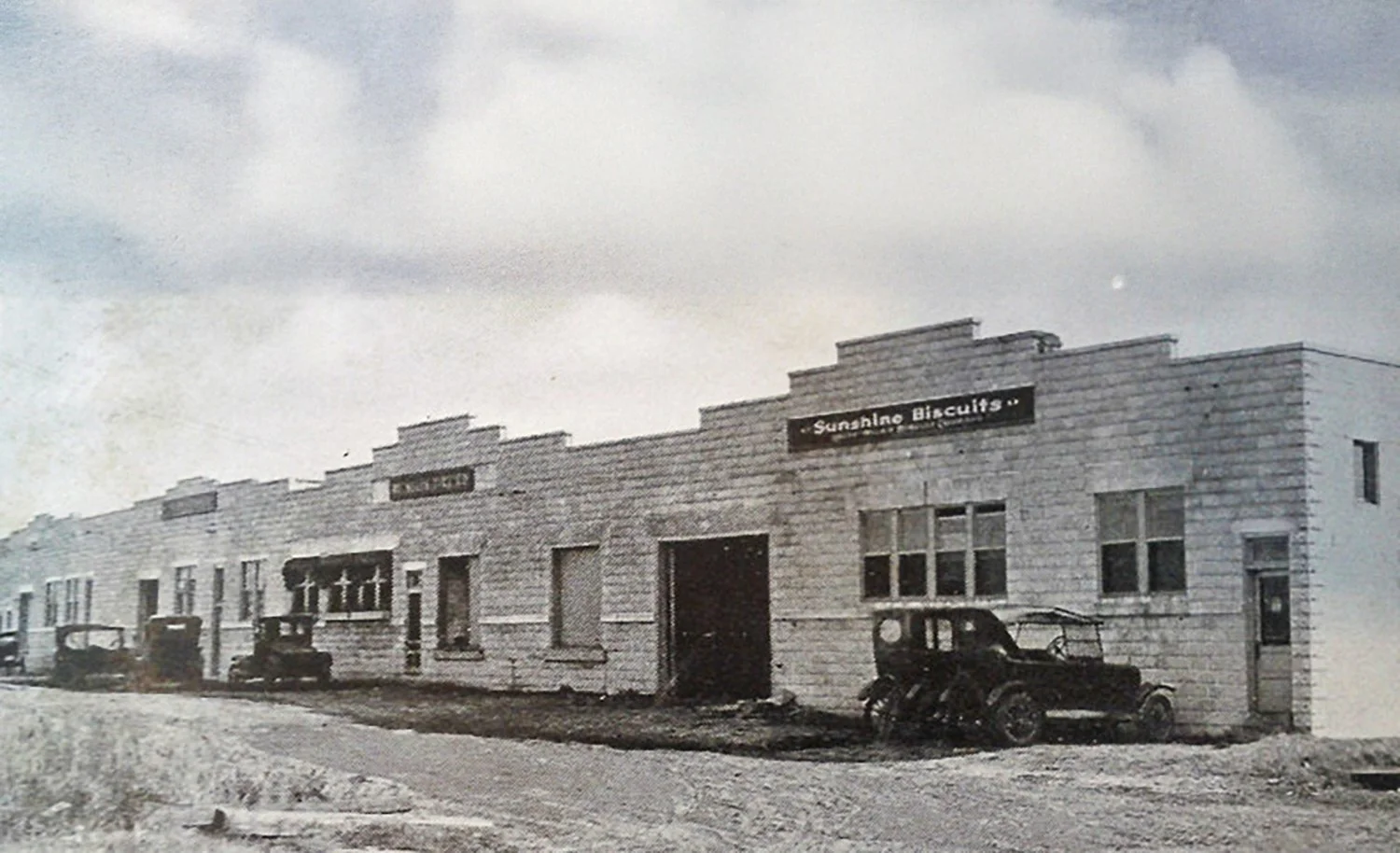 Old black-and-white photo of a row of storefronts, including Sunshine Biscuits, with vintage cars parked in front.