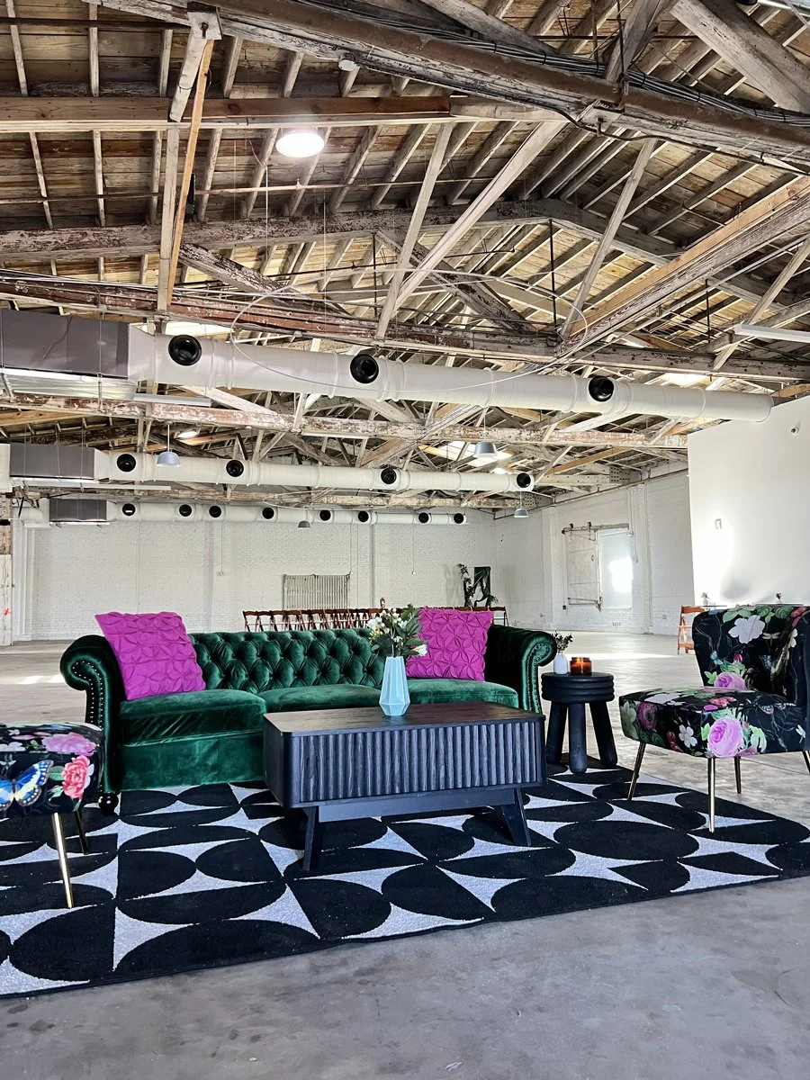 A modern, industrial-style lounge area with a green velvet tufted sofa, colorful floral and pink cushions, patterned chairs, a black and white rug, a black coffee table with a blue vase and flowers, in a spacious room with exposed ceiling beams and ductwork.
