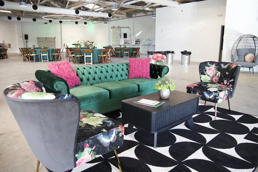 A cozy lounge area with a green tufted velvet sofa, two floral-patterned chairs, and a black coffee table on a black and white geometric rug, with a large open space and tables in the background.