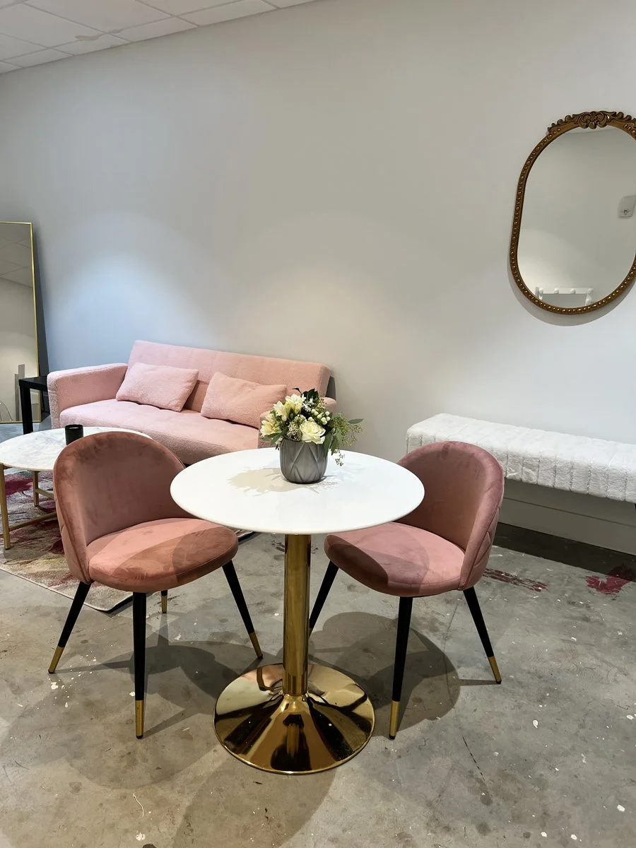 A small white round table with a gold base, two pink velvet chairs, a pink velvet sofa with throw pillows, a vase of white flowers on the table, a white bench underneath a decorative gold mirror, and a wall mirror in the background.