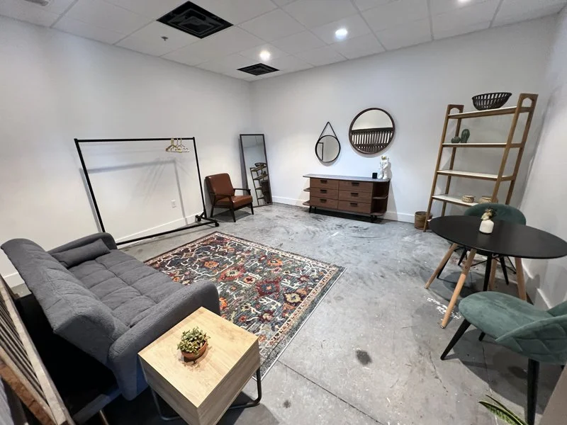 A furnished room with a gray sofa, an area rug, a small side table with a plant, a brown armchair, a tall mirror, a wooden dresser with decorations, a circular mirror, a ladder shelf with decor, a small round black table with two teal chairs, and a white sculpture on the dresser.