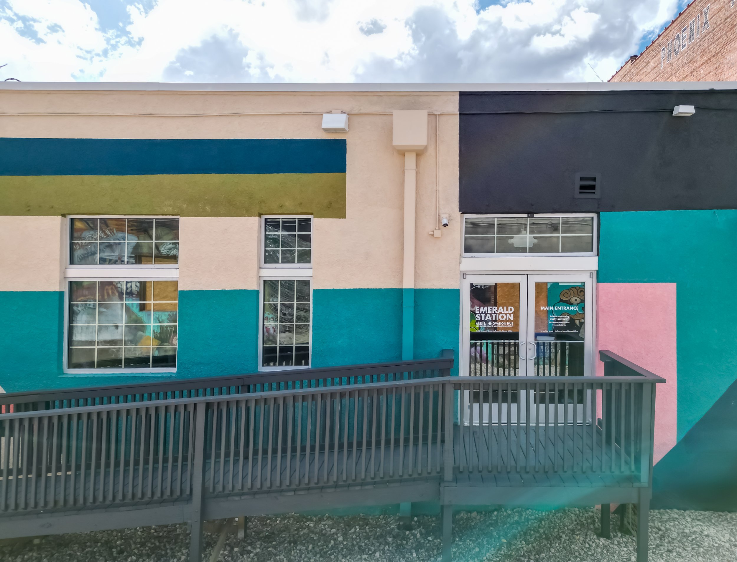 Colorful building with teal, pink, black, and beige sections, featuring large windows, a door labeled 'Emerald Station,' and a wooden ramp outside.