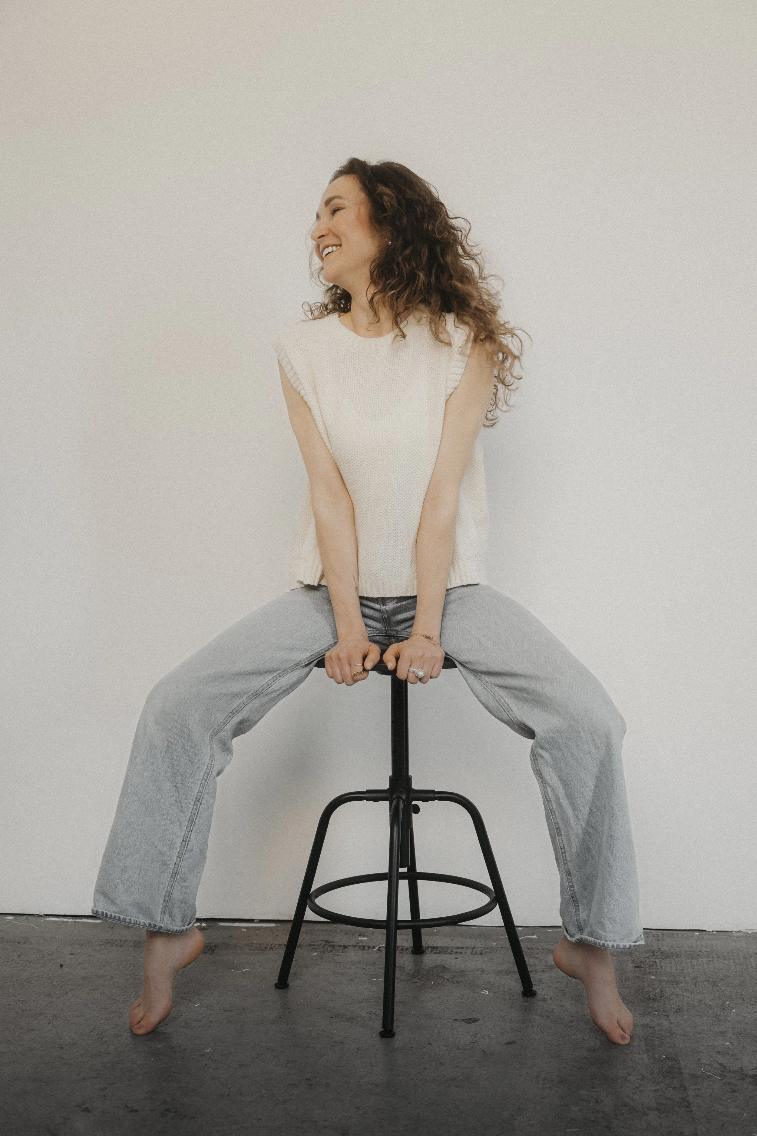 Eine lachende Frau mit lockigen Haaren sitzt auf einem Barhocker gegen eine weiße Wand, trägt eine weiße Strickweste und helle Jeans, barfuß, in einer entspannten Pose.