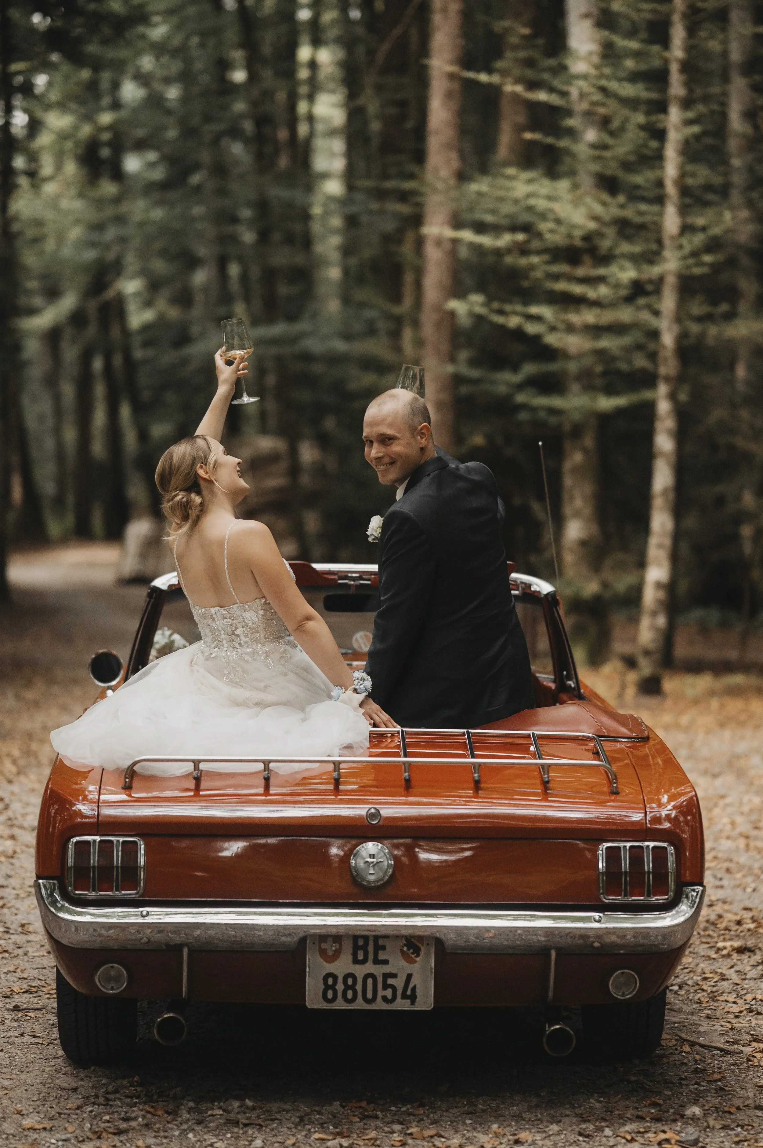 Ein Brautpaar sitzt in einem roten Oldtimer auf einem Waldweg, beide heben mit Champagnergläsern an, in einer schönen Waldkulisse bei Sonnenuntergang, feierliche Atmosphäre, Hochzeitskleidung