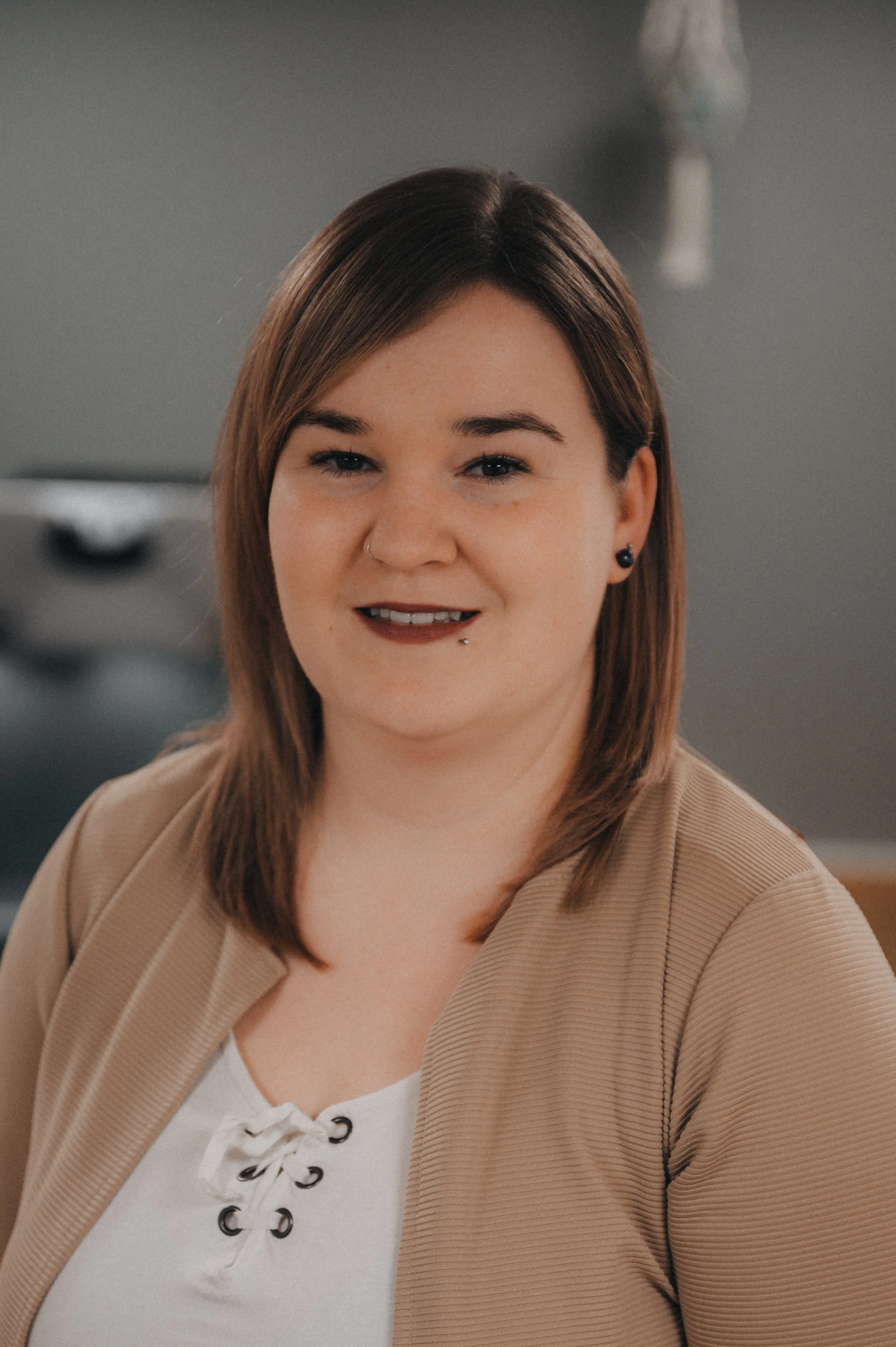 Ein Porträt einer lächelnden Frau mit braunen Haaren, Nasenpiercing und Piercing am Unterlippenrand, trägt eine beige Jacke und ein weißes Top.