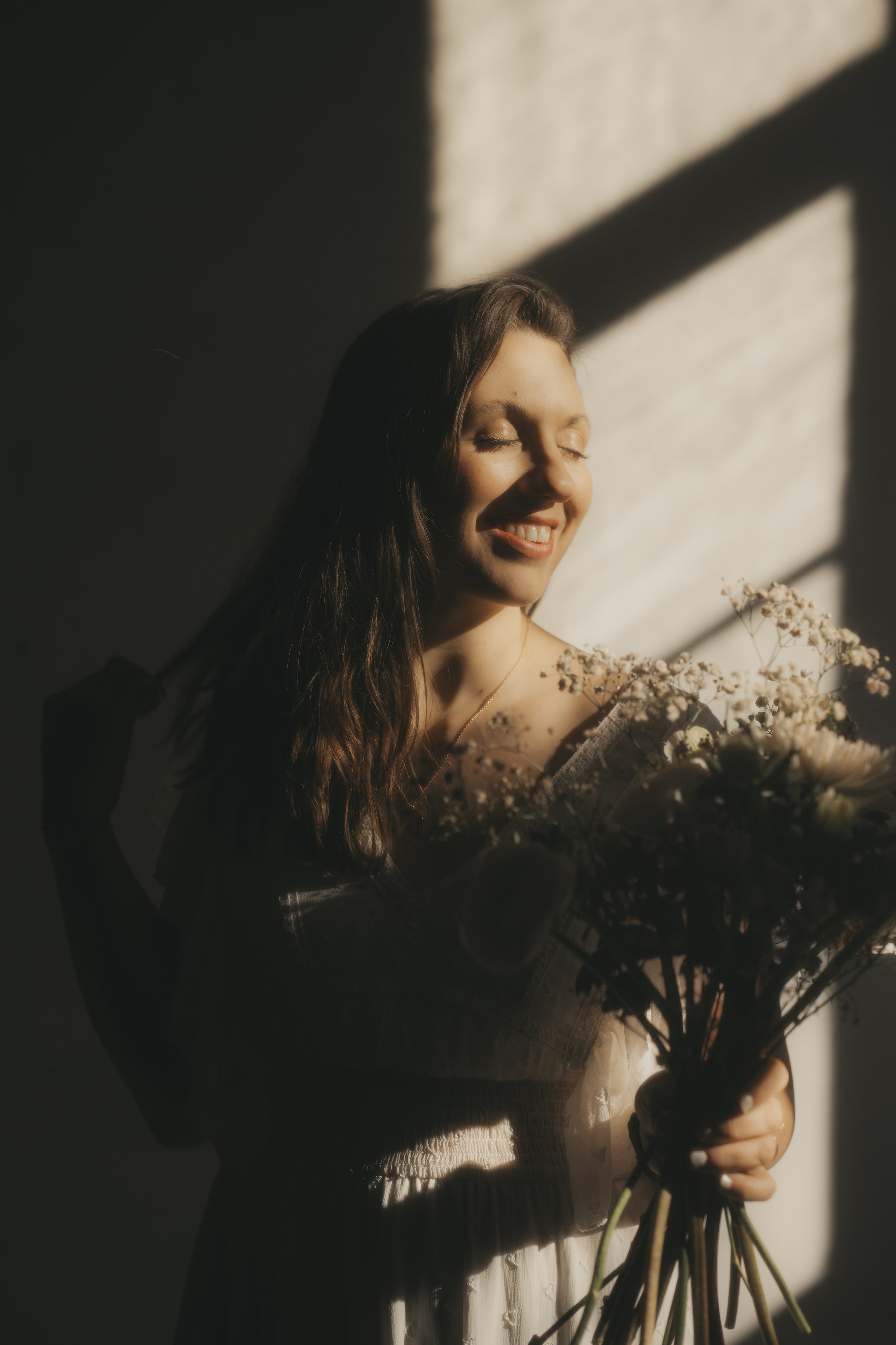 Eine lächelnde Frau mit geschlossenen Augen, die einen Blumenstrauß in den Händen hält, im warmen Sonnenlicht, mit Schatten an der Wand im Hintergrund.