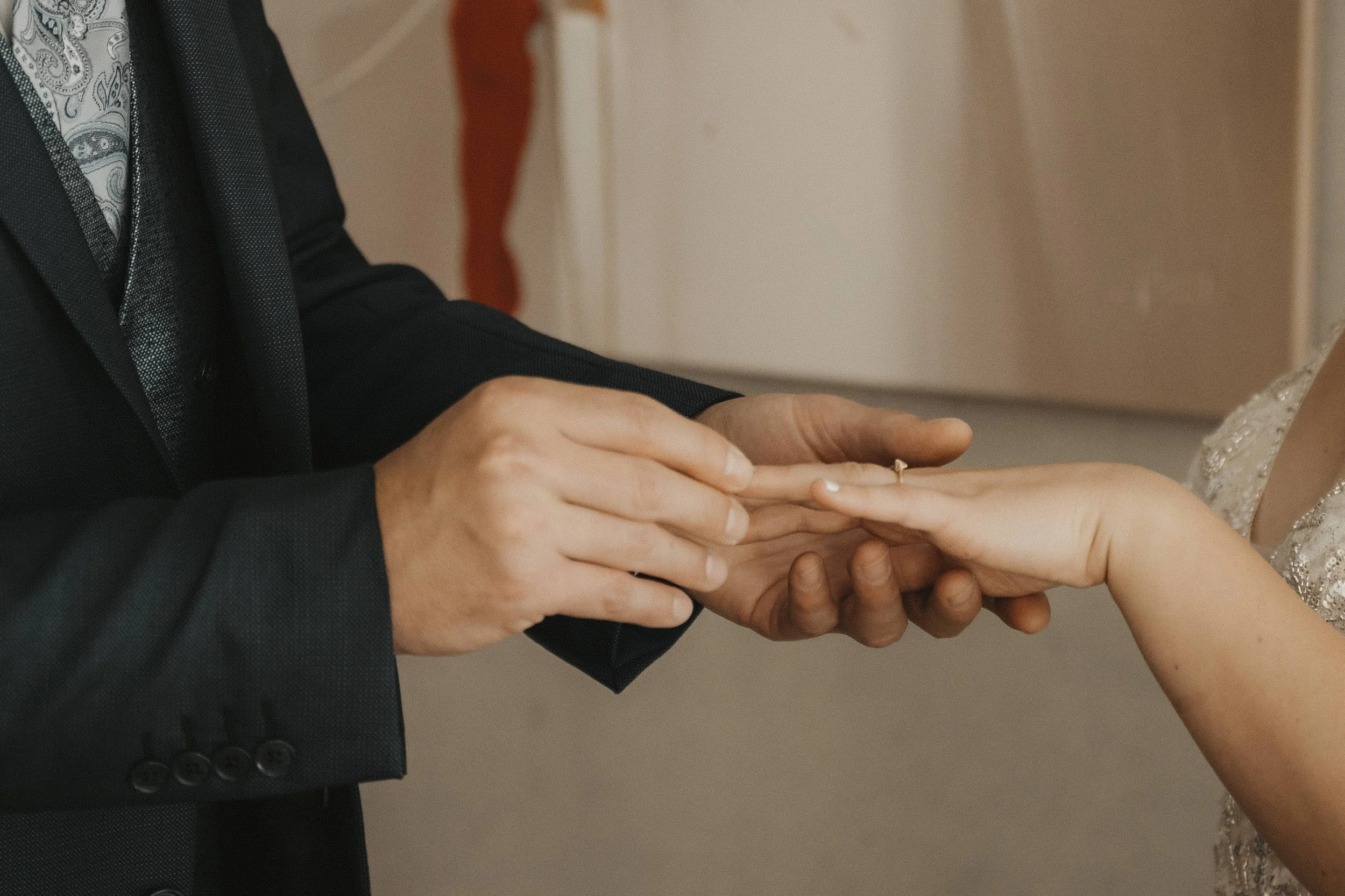 Ein Mann in einem schwarzen Anzug hält die Hand einer Frau, vermutlich während einer Hochzeitszeremonie, und steckt ihr den Ehering an.