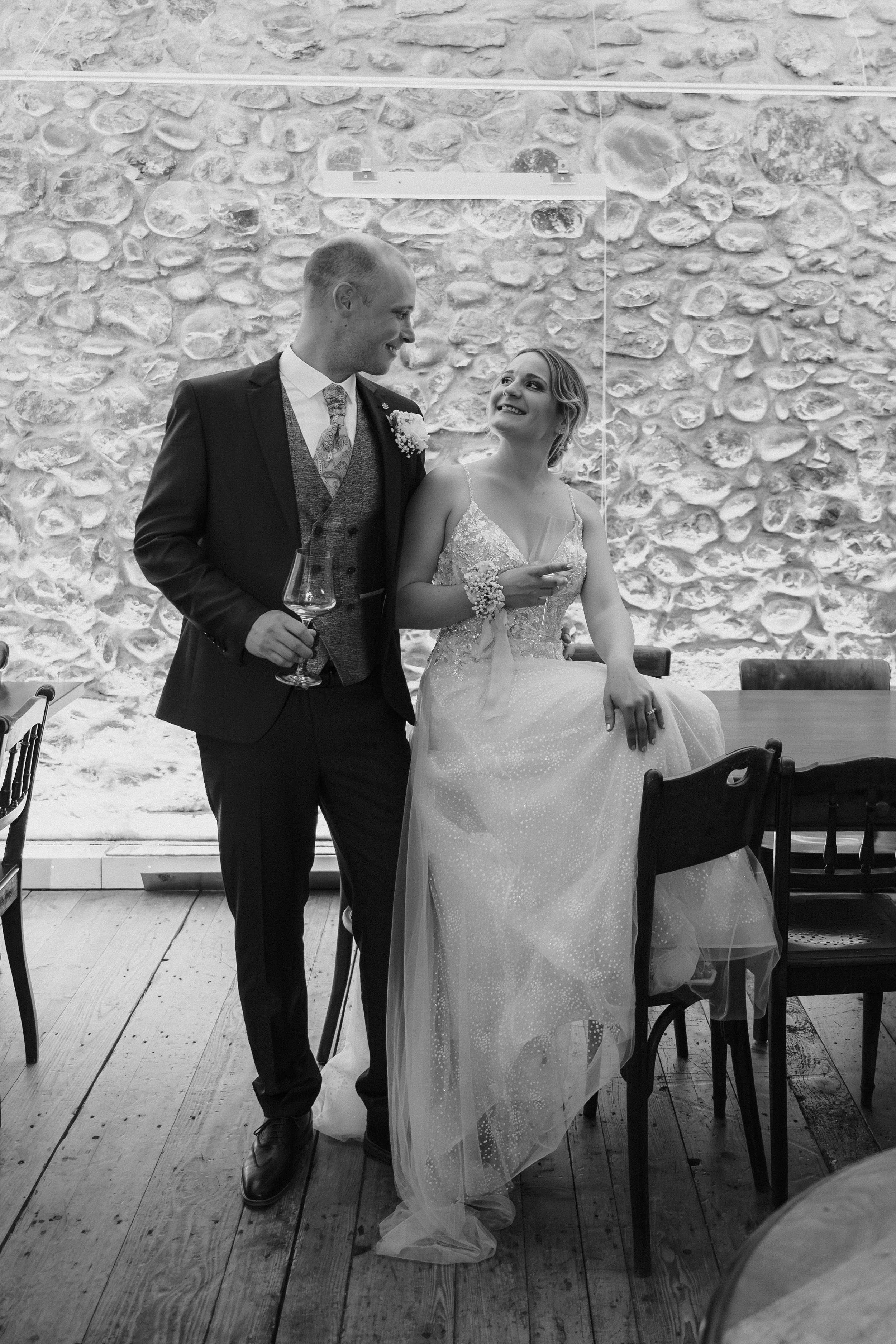 Ein Paar bei einer Hochzeitsfeier, der Bräutigam mit einem Glas Wein und die Braut, die auf einem Stuhl sitzt, lachen und schauen sich an, im Hintergrund eine Steinmauer.
