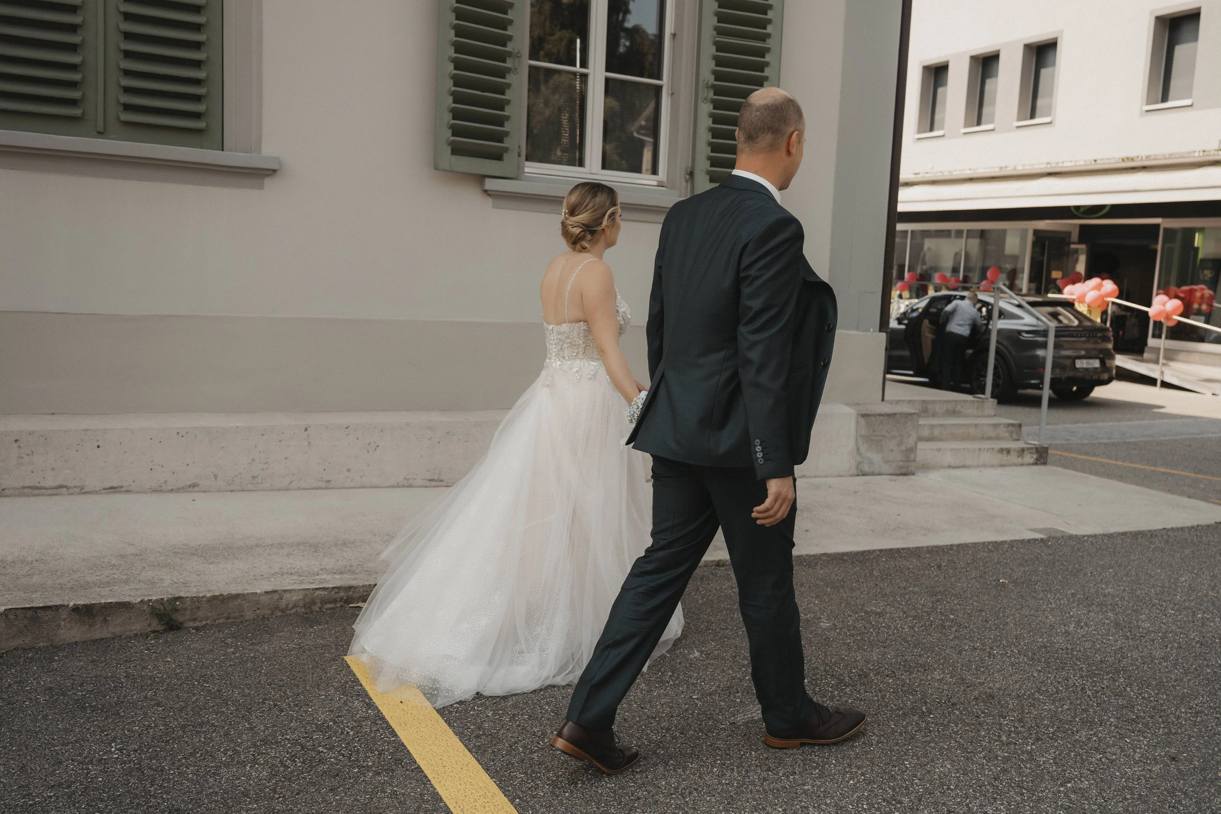 Ein Brautpaar geht getrennt, die Braut trägt ein weißes Hochzeitskleid und die Figur mit Anzug ist ein Mann, vermutlich der Bräutigam, vor einem Gebäude in einer Stadtumgebung.