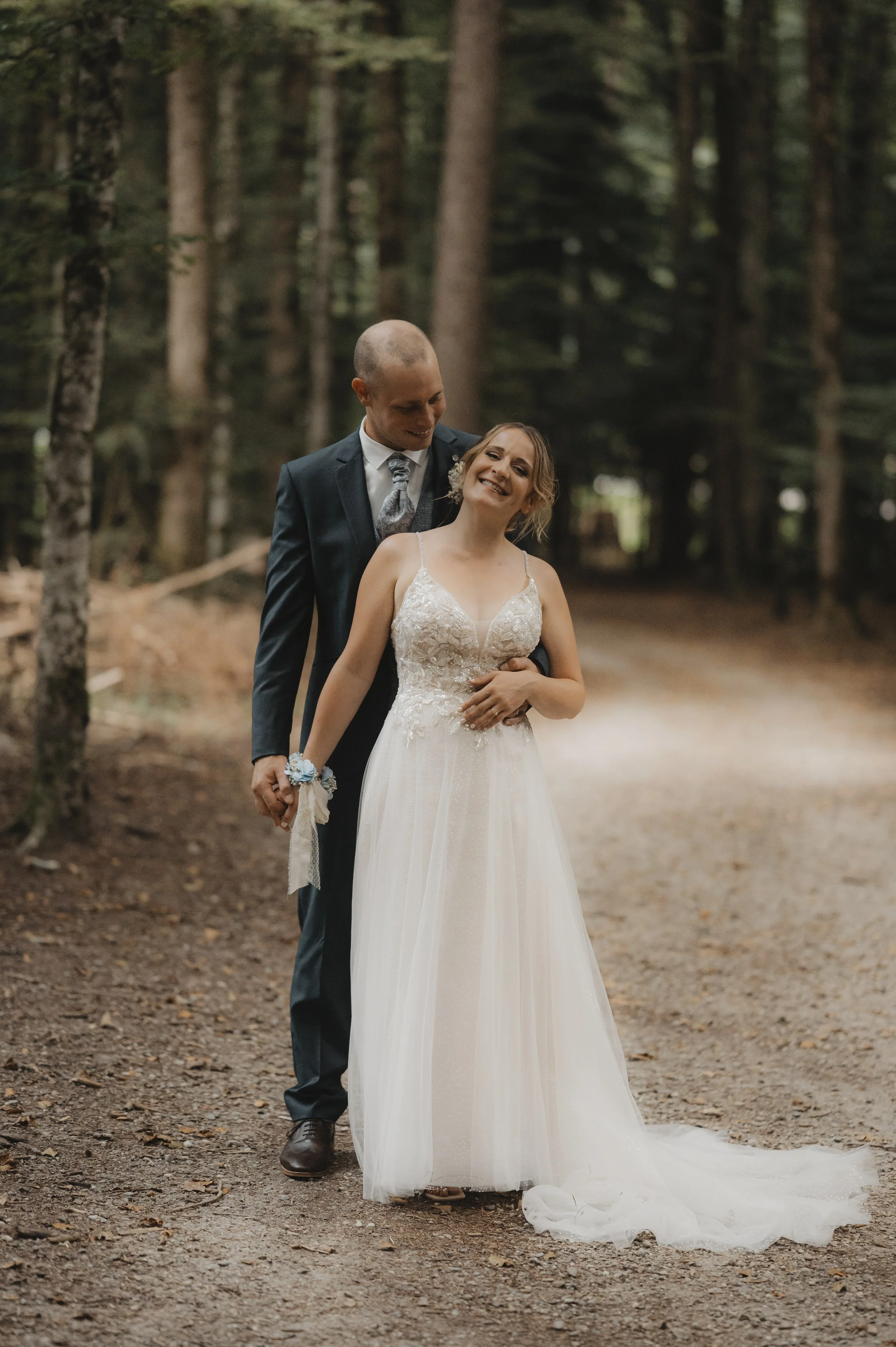 Ein Brautpaar in formeller Kleidung, die durch einen Wald spaziereng auf einer Wege aus Erd und Blättern.
