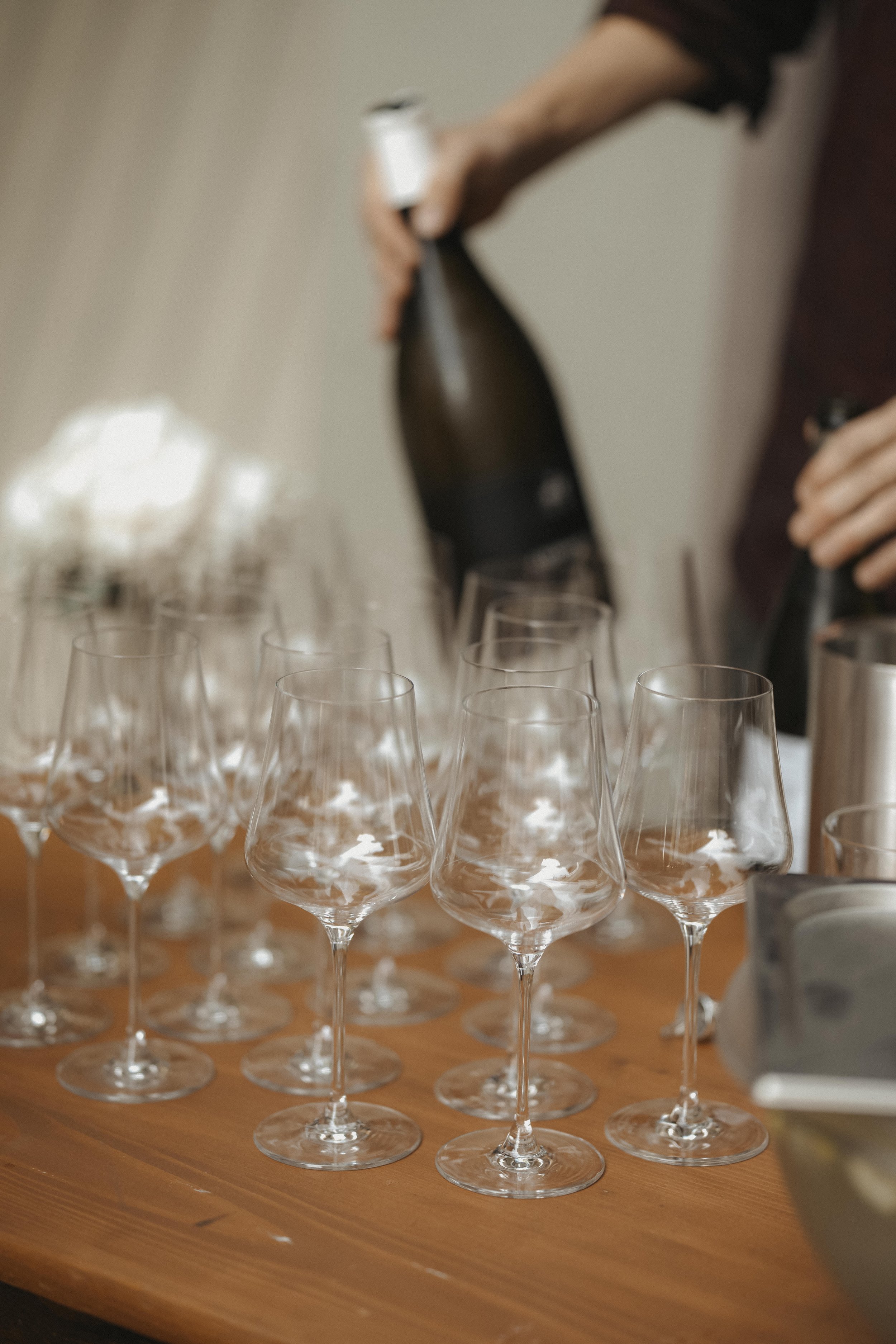 Mehrere leere Weingläser auf einem Holztisch, im Hintergrund eine Person, die eine Flasche Wein einschenkt.