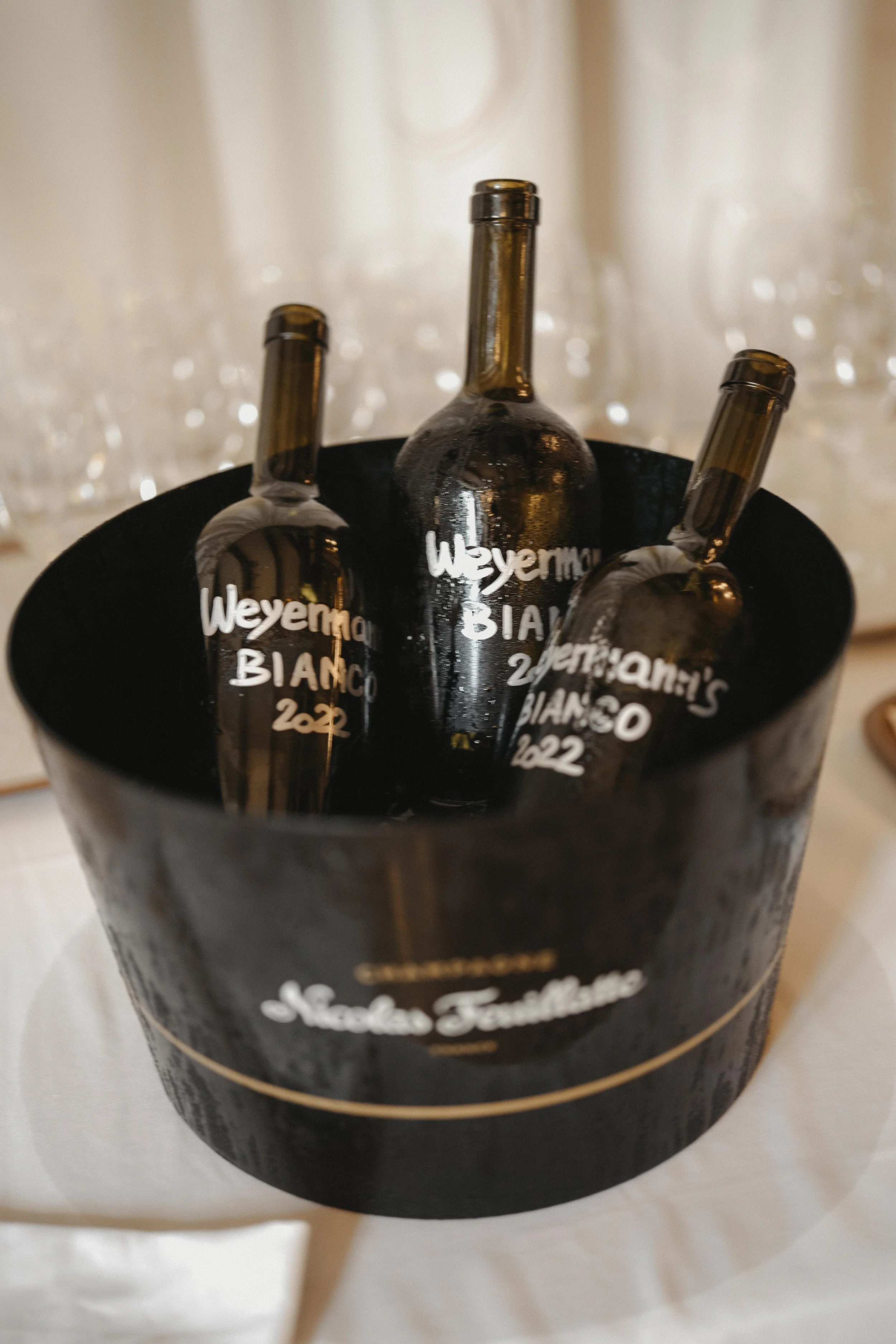 Drei Weinflaschen in einem schwarzen Eimer, beschriftet mit 'Weyenmaier BIANCO 2022', auf einem weißen Tisch.