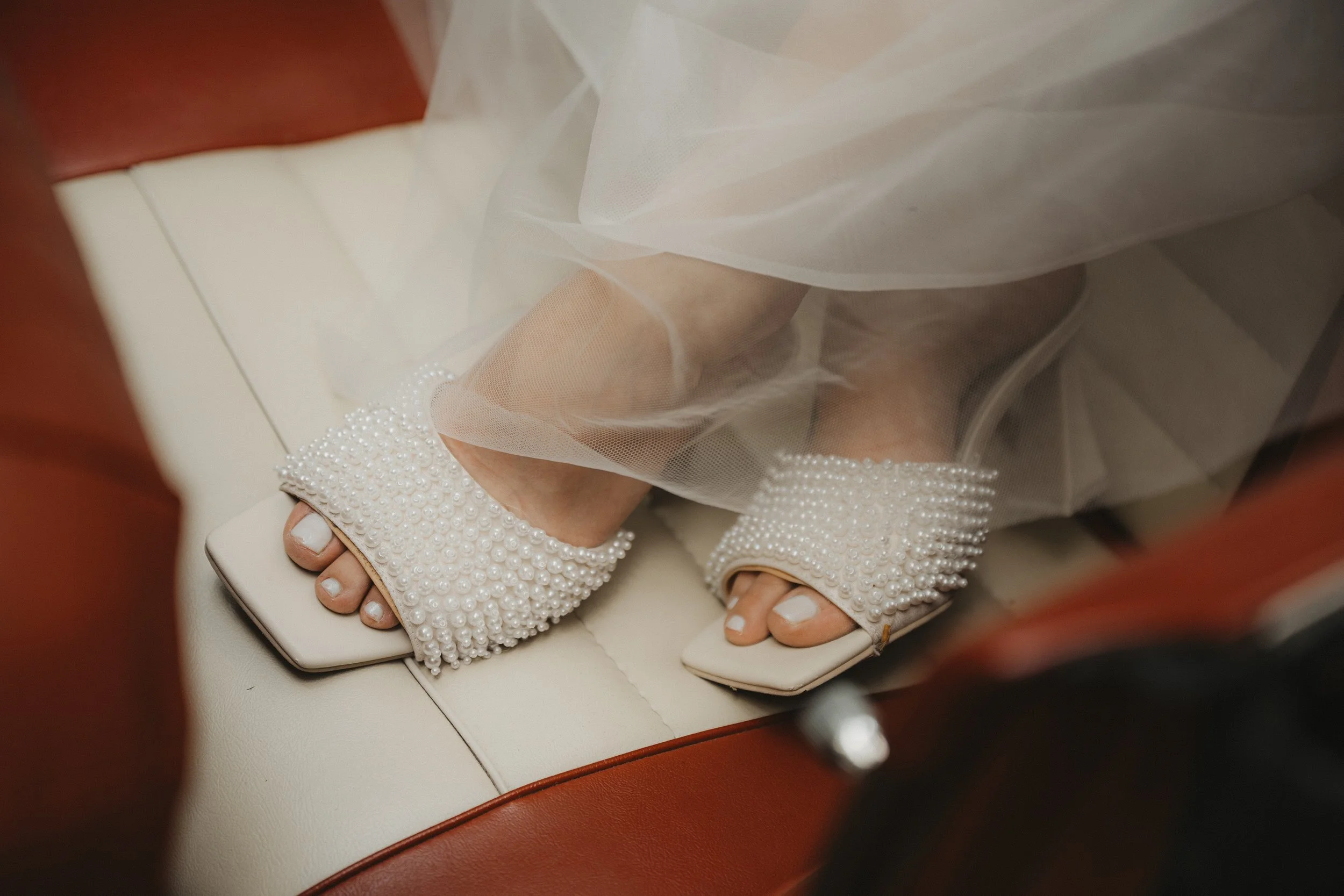 Nahaufnahme der Beine einer Frau, die auf einem weißen Polstersitz sitzt, trägt elegante Schuhe mit Perlenverzierung und eine Tüll- oder Organza-Schleier