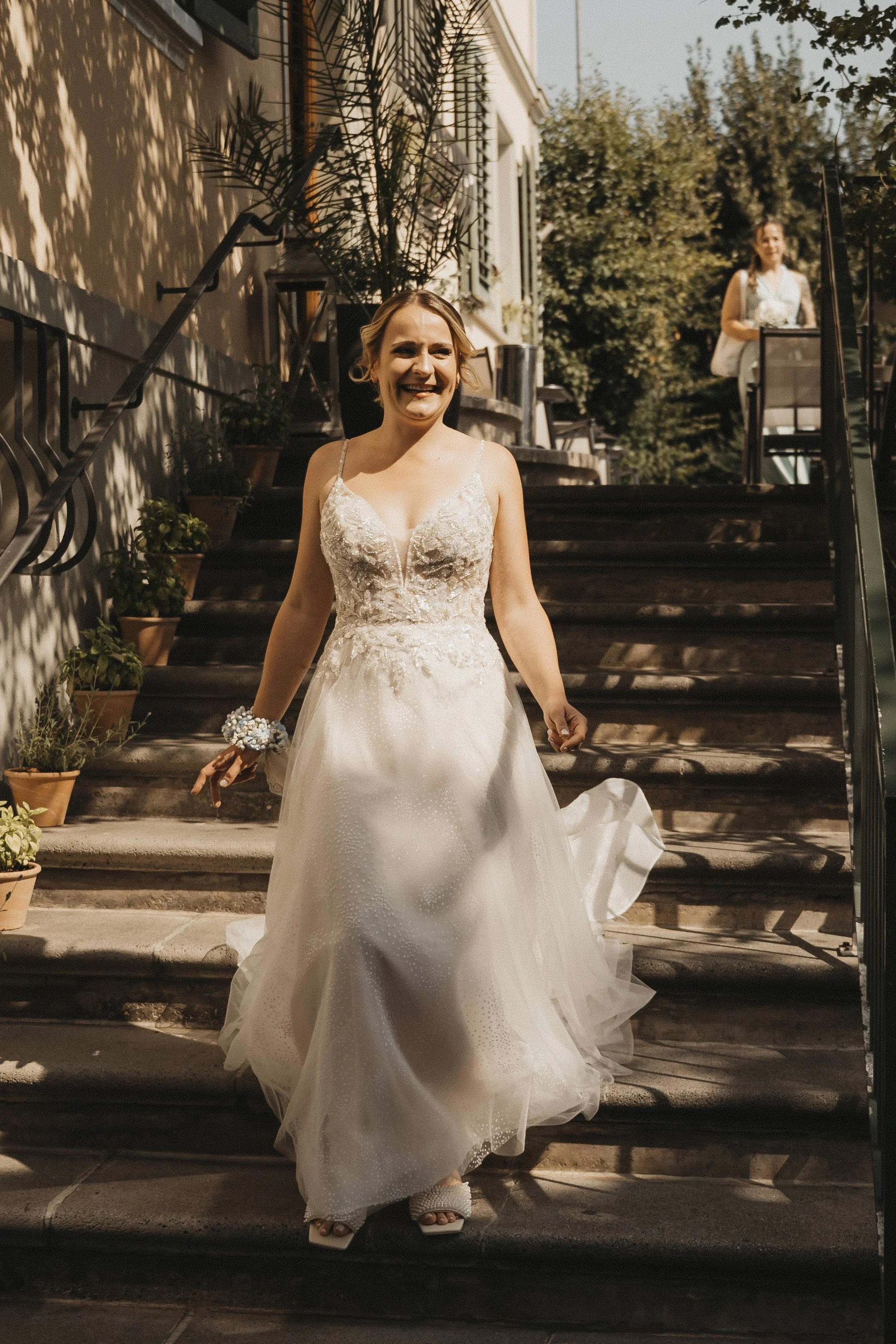 Frau in Brautkleid auf Treppen vor einem Gebäude, lächelnd, mit Pflanzen umgeben, im Freien an sonnigem Tag.