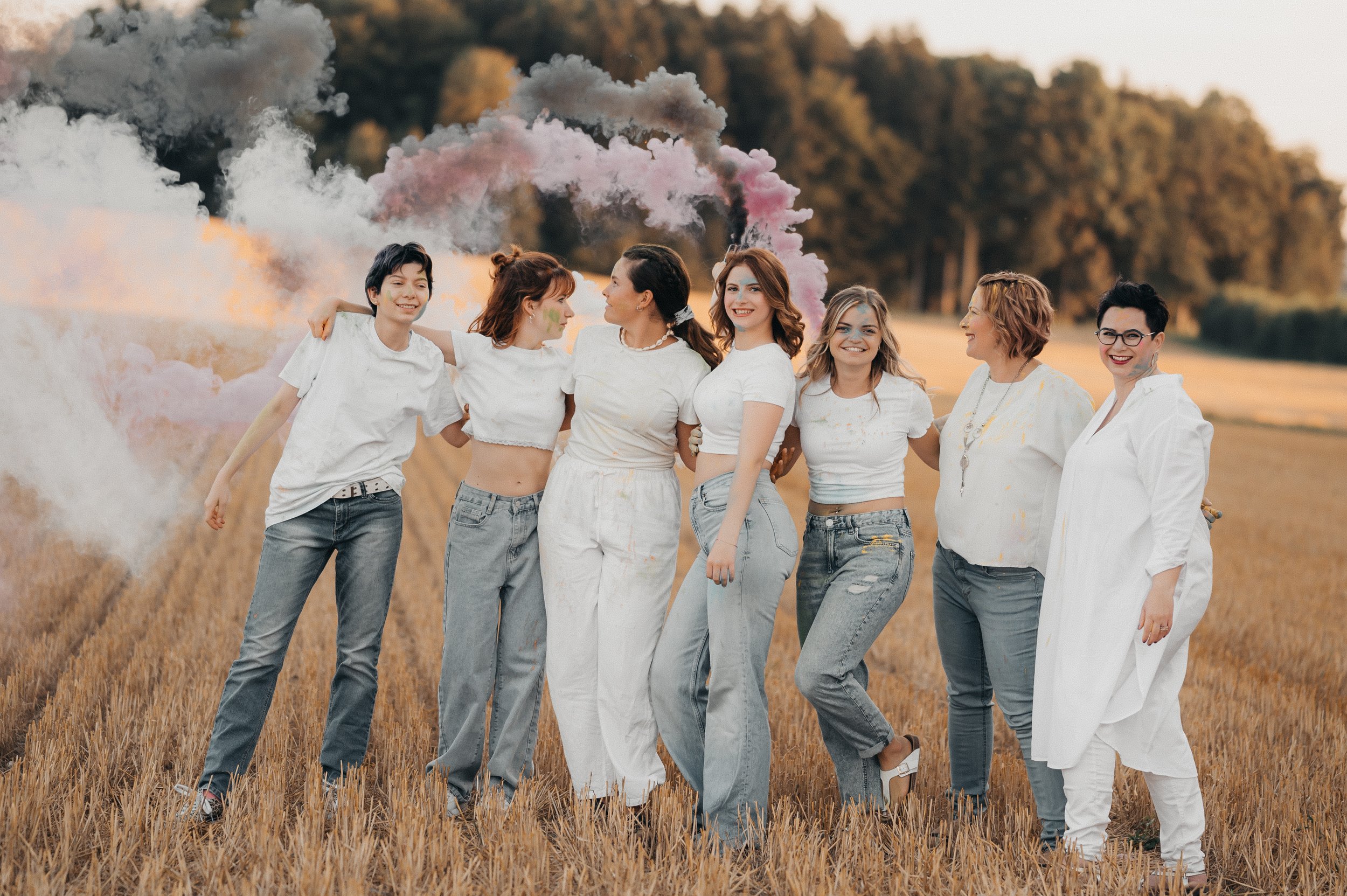 Eine Gruppe von acht Frauen in einem Feld, die gemeinsam pastellfarbene Rauchfahnen hinter sich aufsteigen lassen, bei Sonnenuntergang, lachend und in weißen Kleidung.