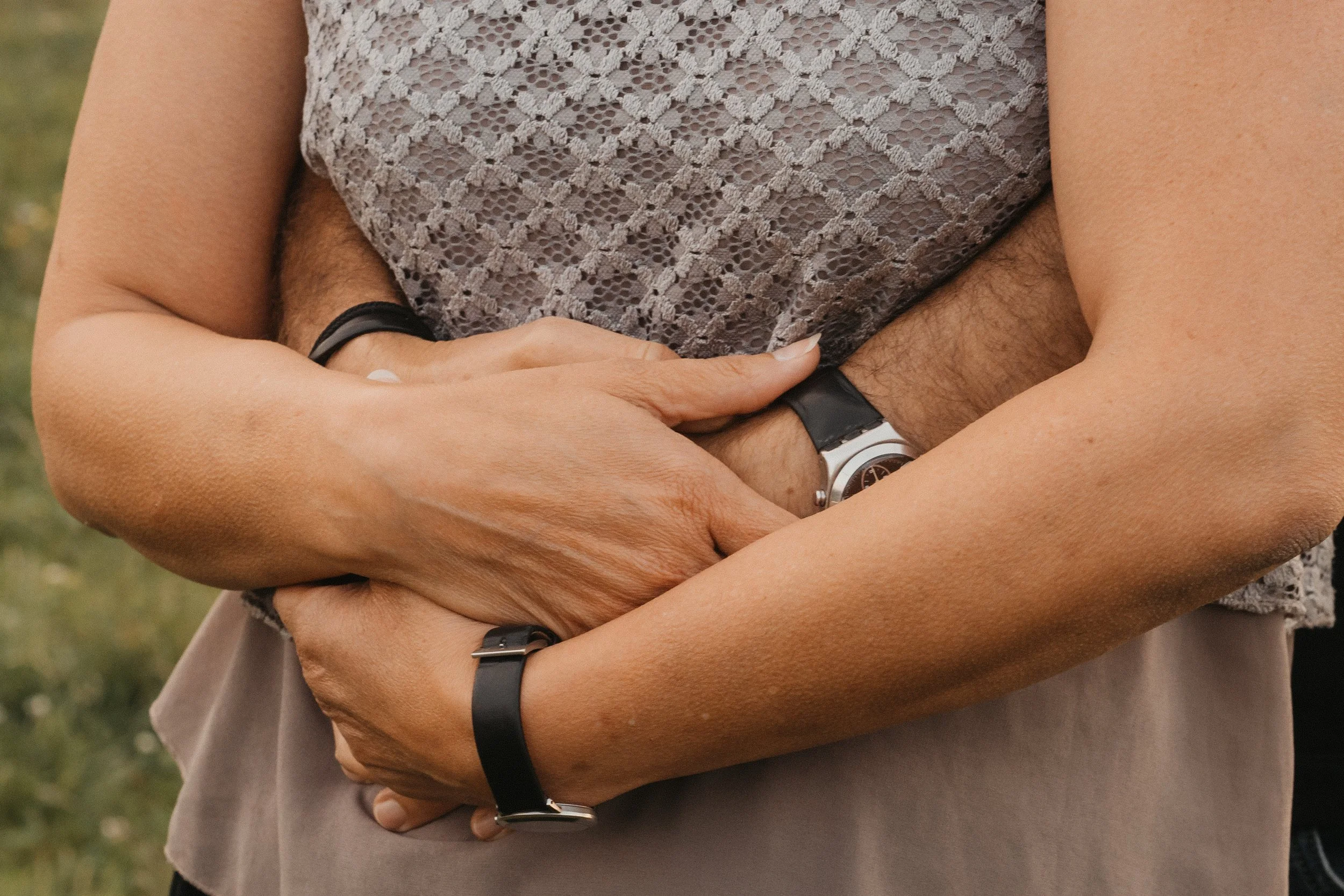 Nahaufnahme von zwei Personen, die sich umarmen, mit sichtbaren Armen und Händen, beide tragen Uhren und Armbänder, im Hintergrund eine grüne Wiese.