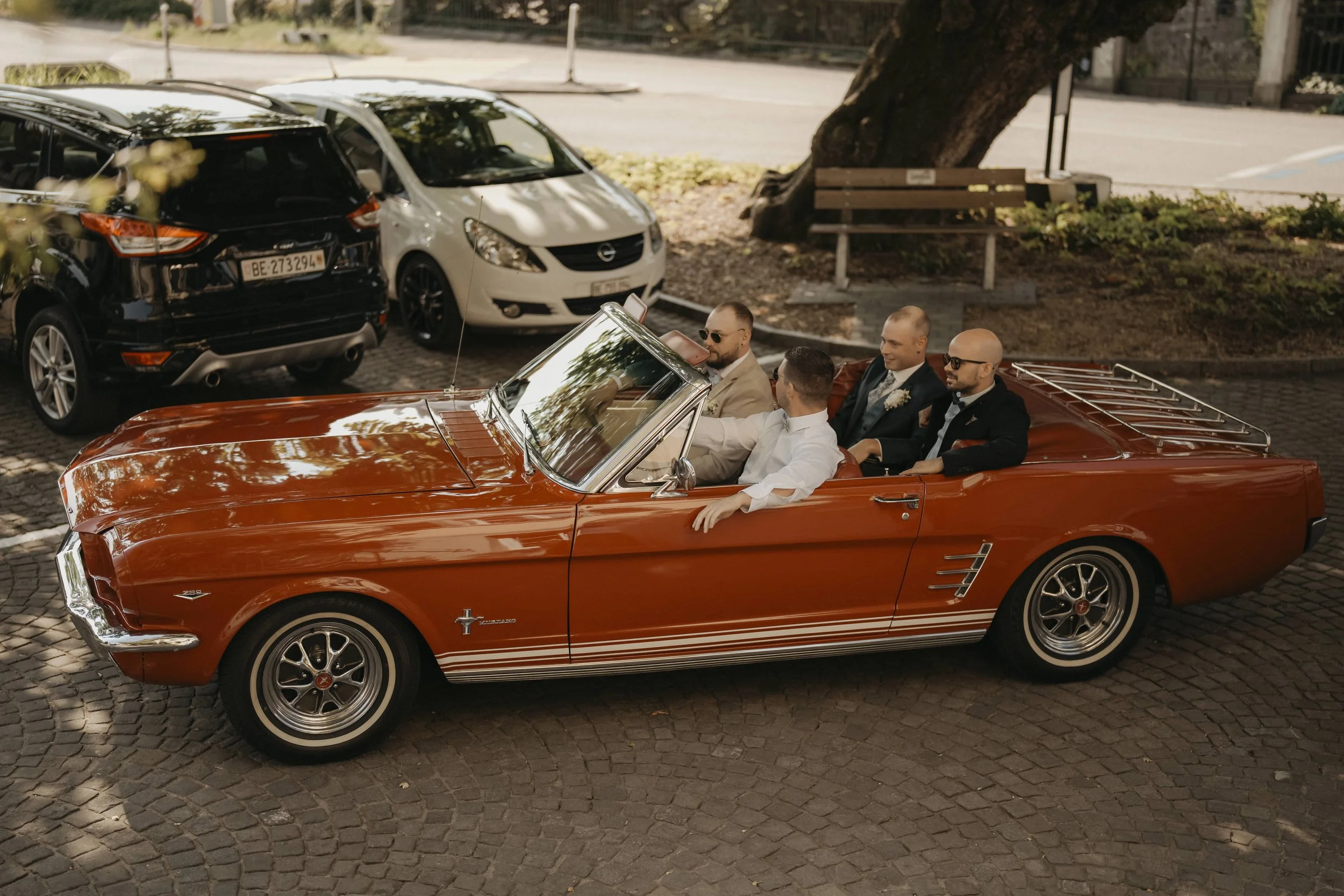 Fünf Männer sitzen in einem alten orangefarbenen Ford Mustang Cabrio auf einem gepflasterten Parkplatz, umgeben von parkenden Autos und Bäumen.